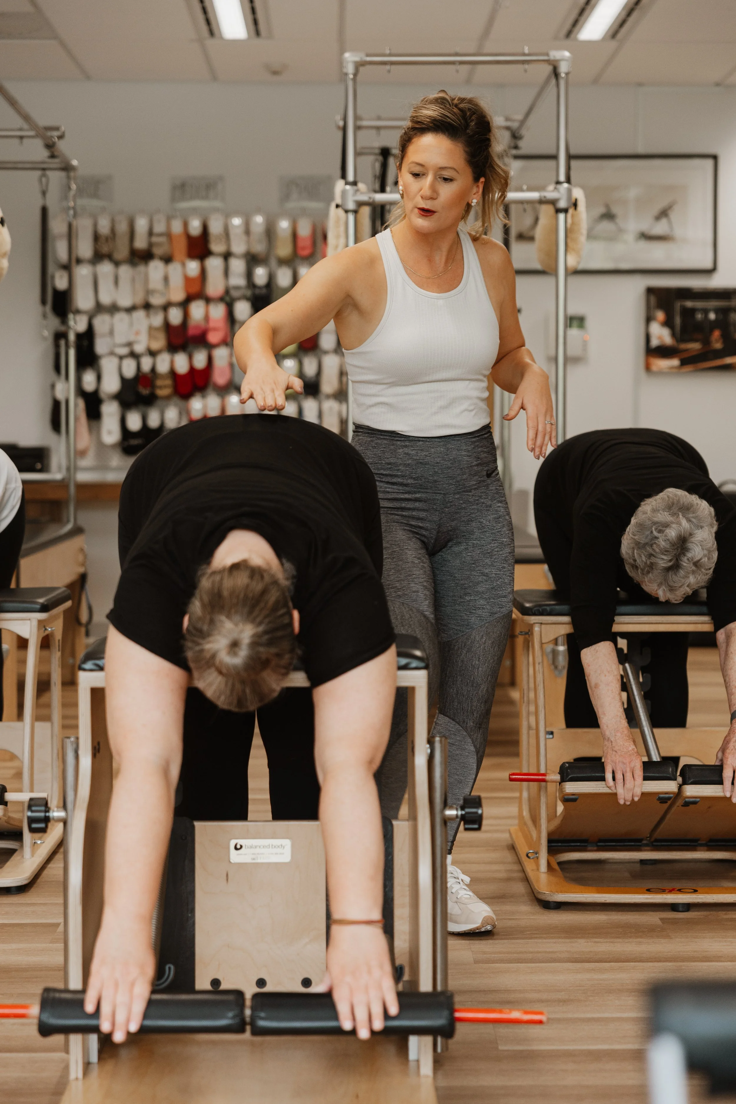 A woman instructs elderly women in a Pilates class at a fitness studio, demonstrating an exercise with Pilates reformers.