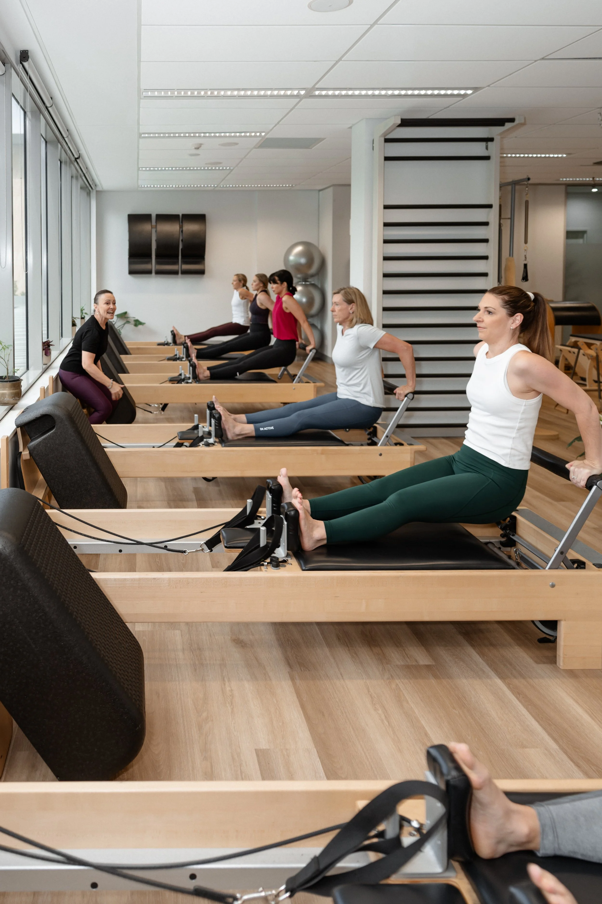 Women participating in a Pilates class using reformer machines in a bright, modern gym with large windows.