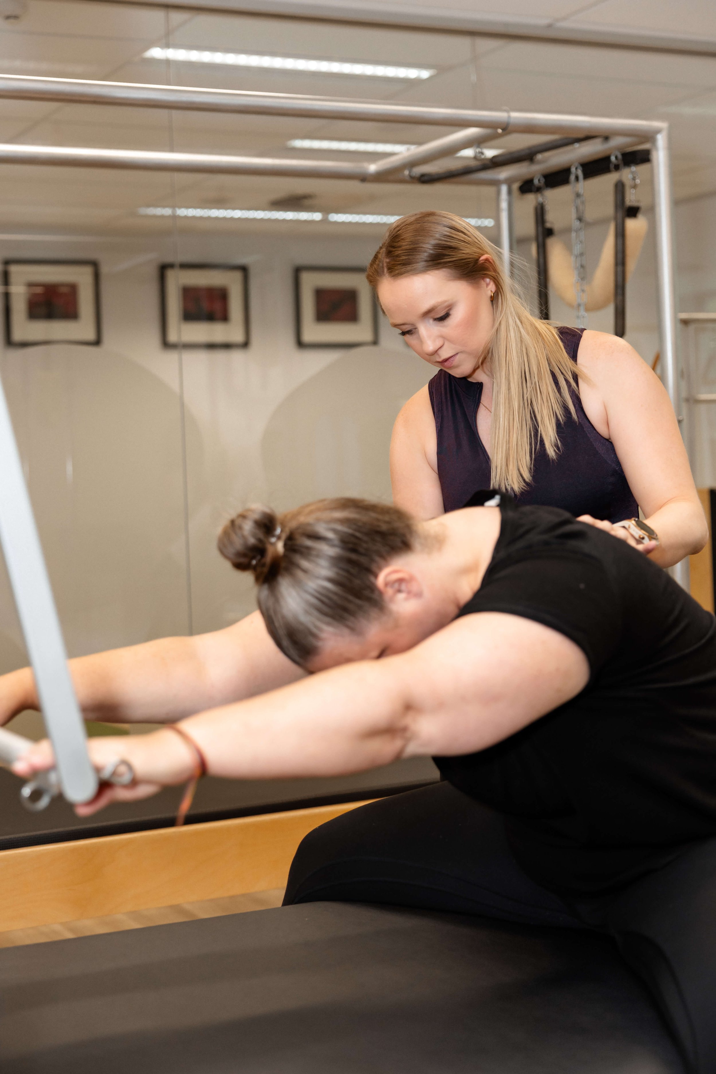 A woman stretching on a Pilates reformer while an instructor observes in a fitness studio.