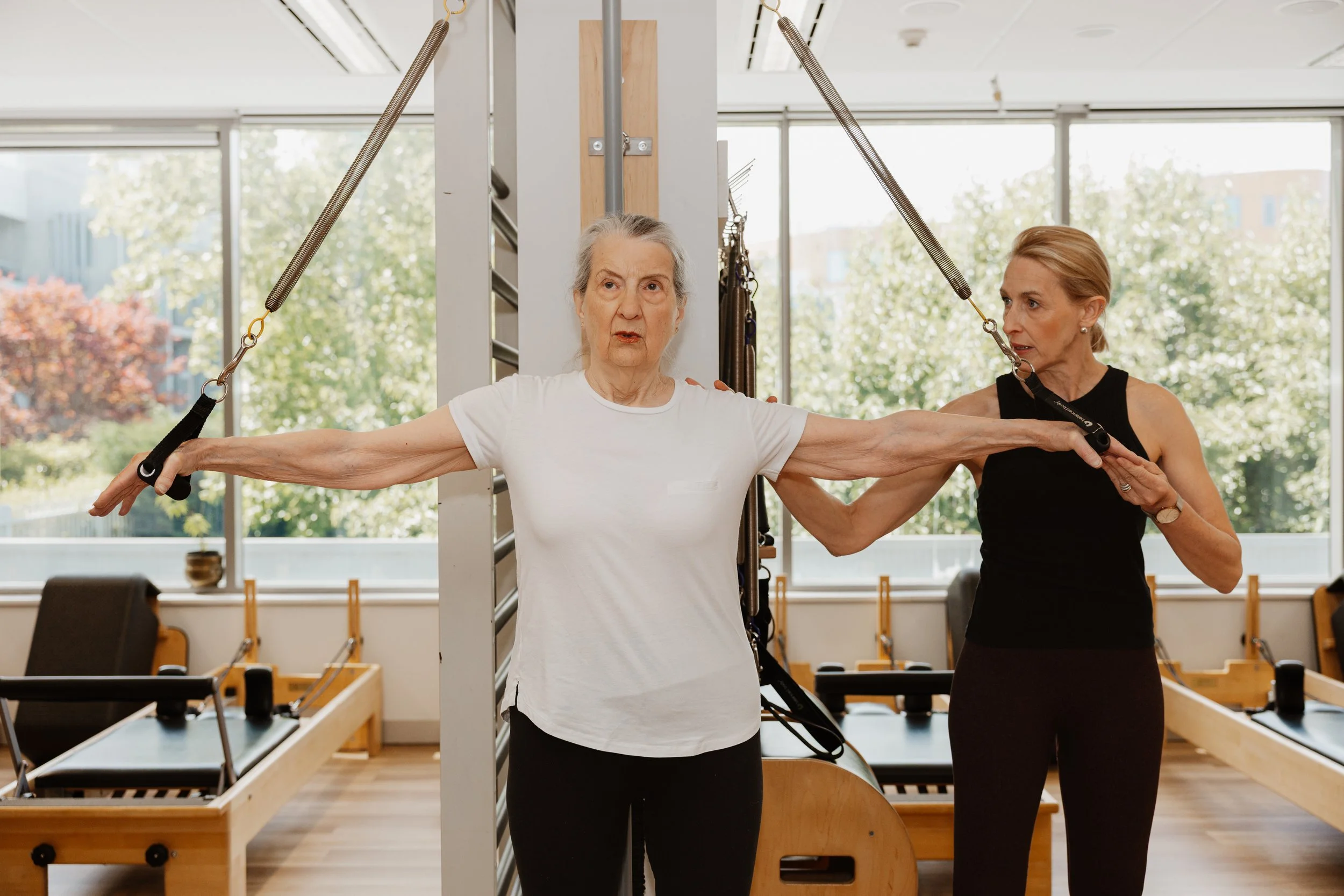 An elderly woman participating in a Pilates exercise session with assistance from an instructor. She is performing an arm stretch using resistance straps in a bright, modern studio.