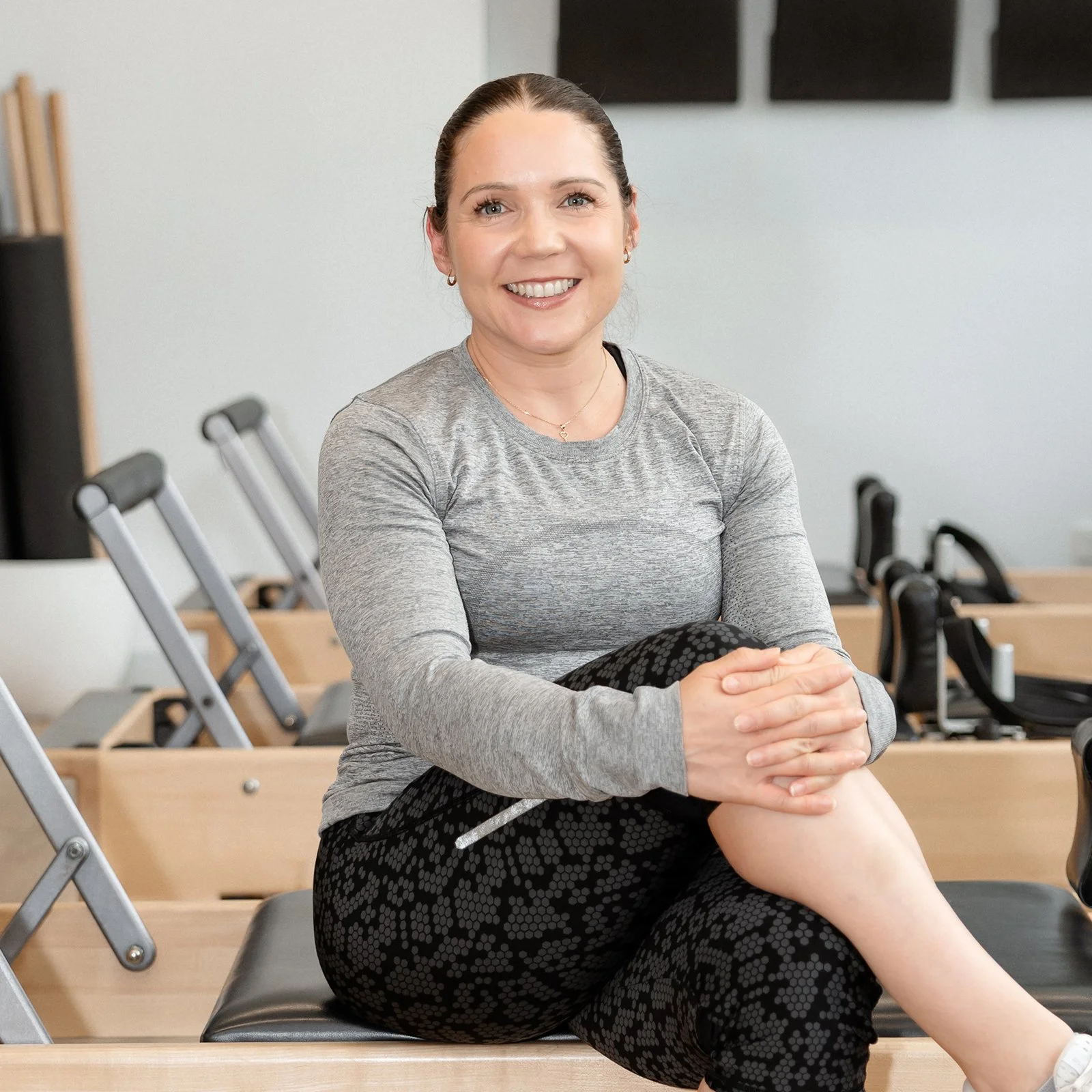 A woman sitting on a black bench in a room with folding chairs. She is smiling, wearing a gray long-sleeve shirt and black patterned leggings, holding her leg with both hands.