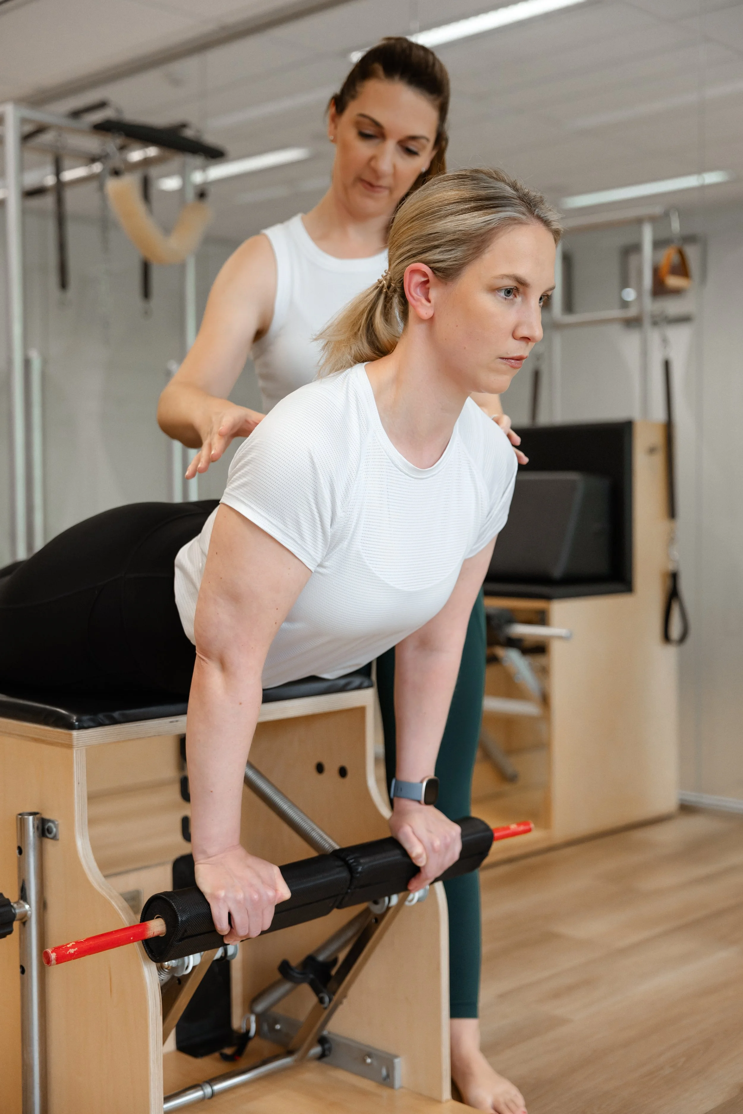 A woman performing a back extension exercise on a Pilates equipment, supervised by a trainer in a gym setting.