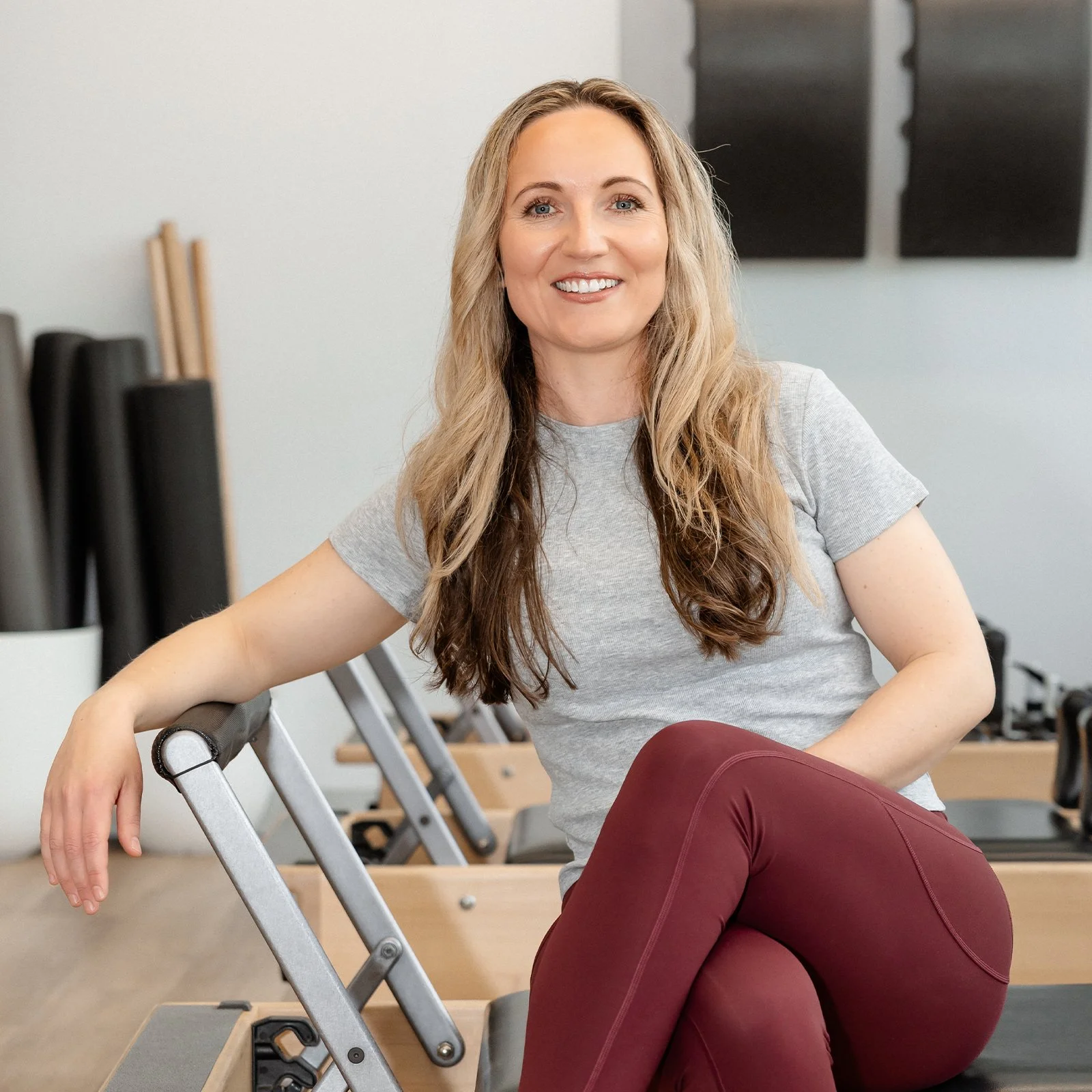 A woman with long wavy hair sitting on a Pilates reformer machine in a fitness studio, smiling at the camera.