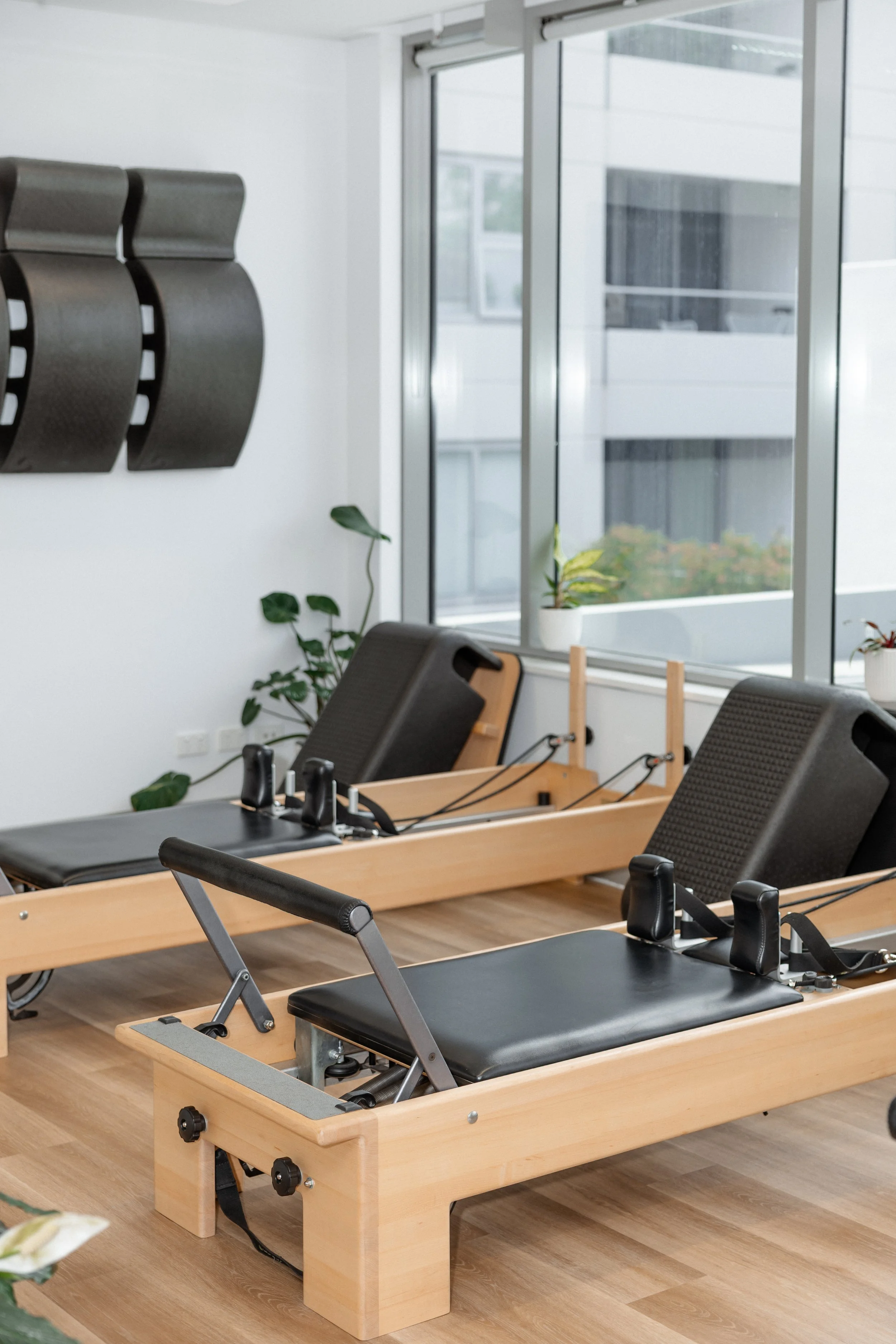 Two Reformer Pilates machines with black padding in a bright studio with large windows and indoor plants.