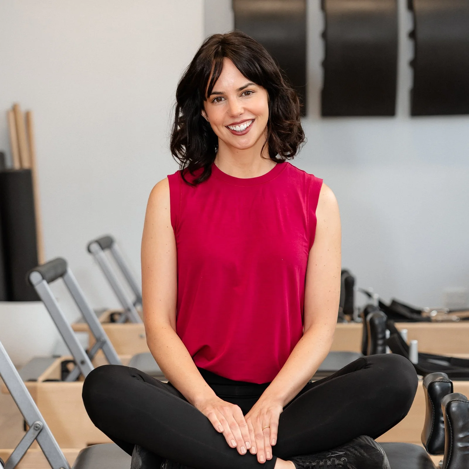 A woman with dark, wavy hair wearing a sleeveless red top and black pants, sitting cross-legged on a bench in a room with exercise equipment, smiling at the camera.