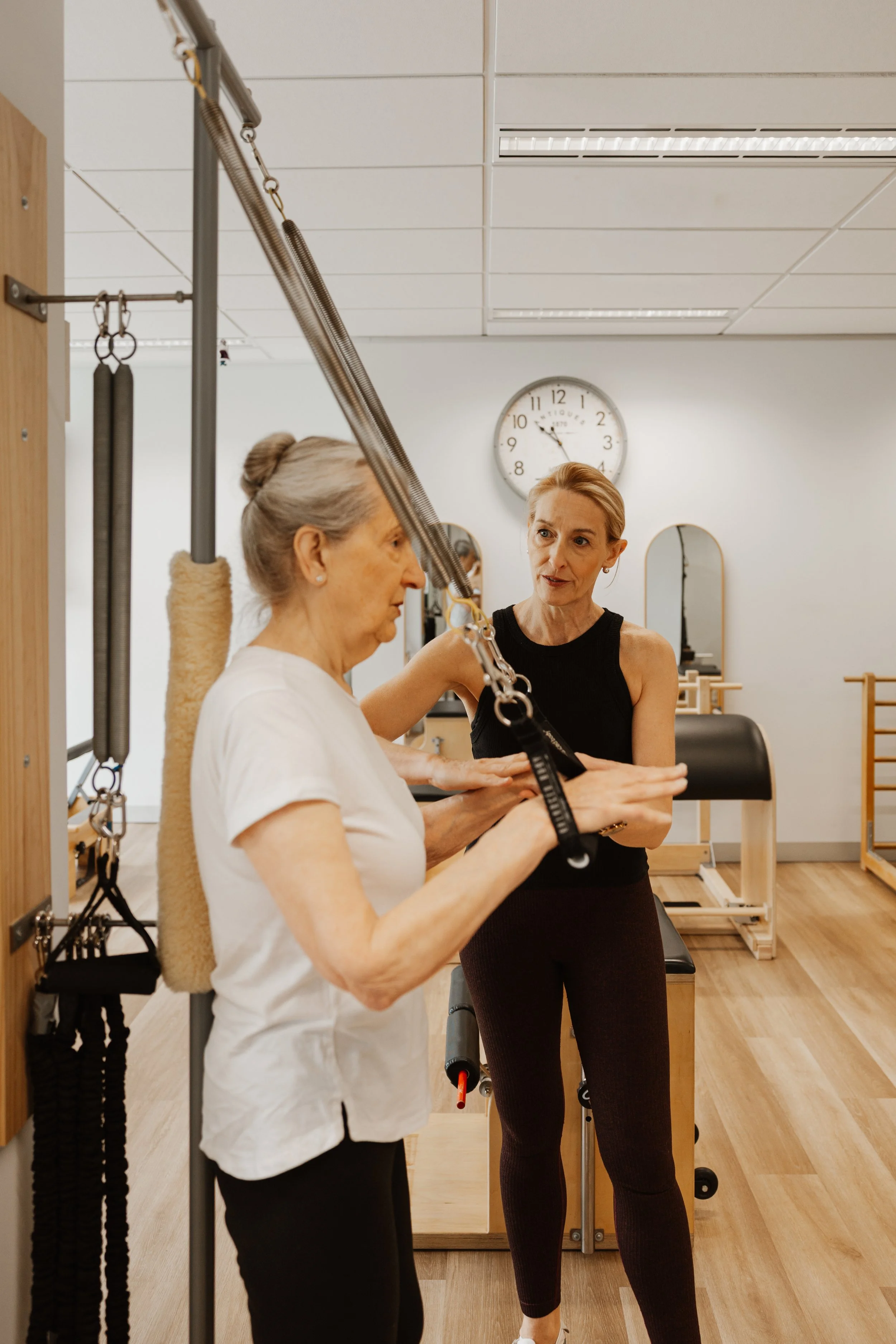 An elderly woman exercises with a suspension trainer under the guidance of a female instructor in a physical therapy or fitness studio.