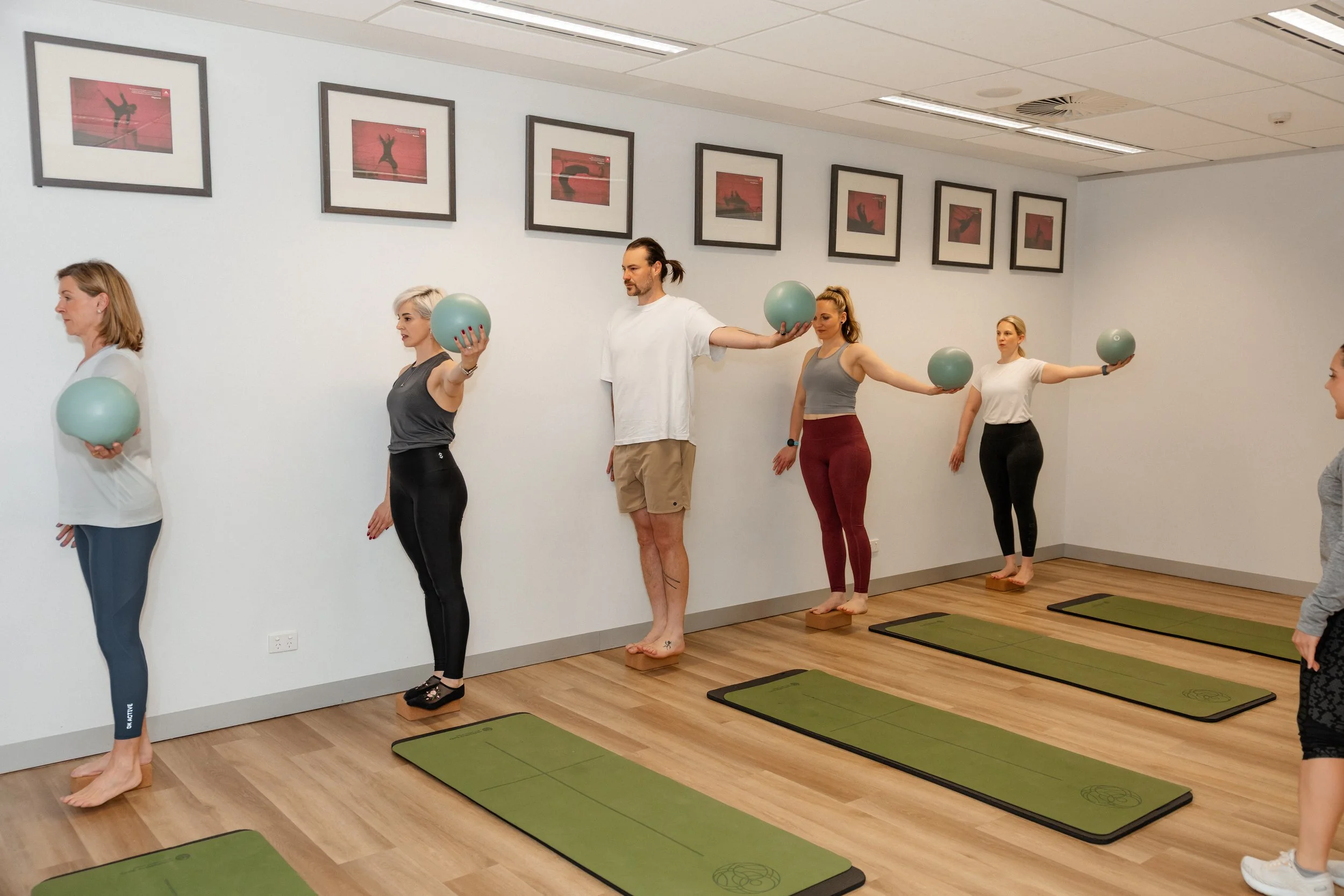 A group of six people participating in a fitness class or exercise session in a studio, standing on green mats, holding exercise balls with their arms extended to the sides.