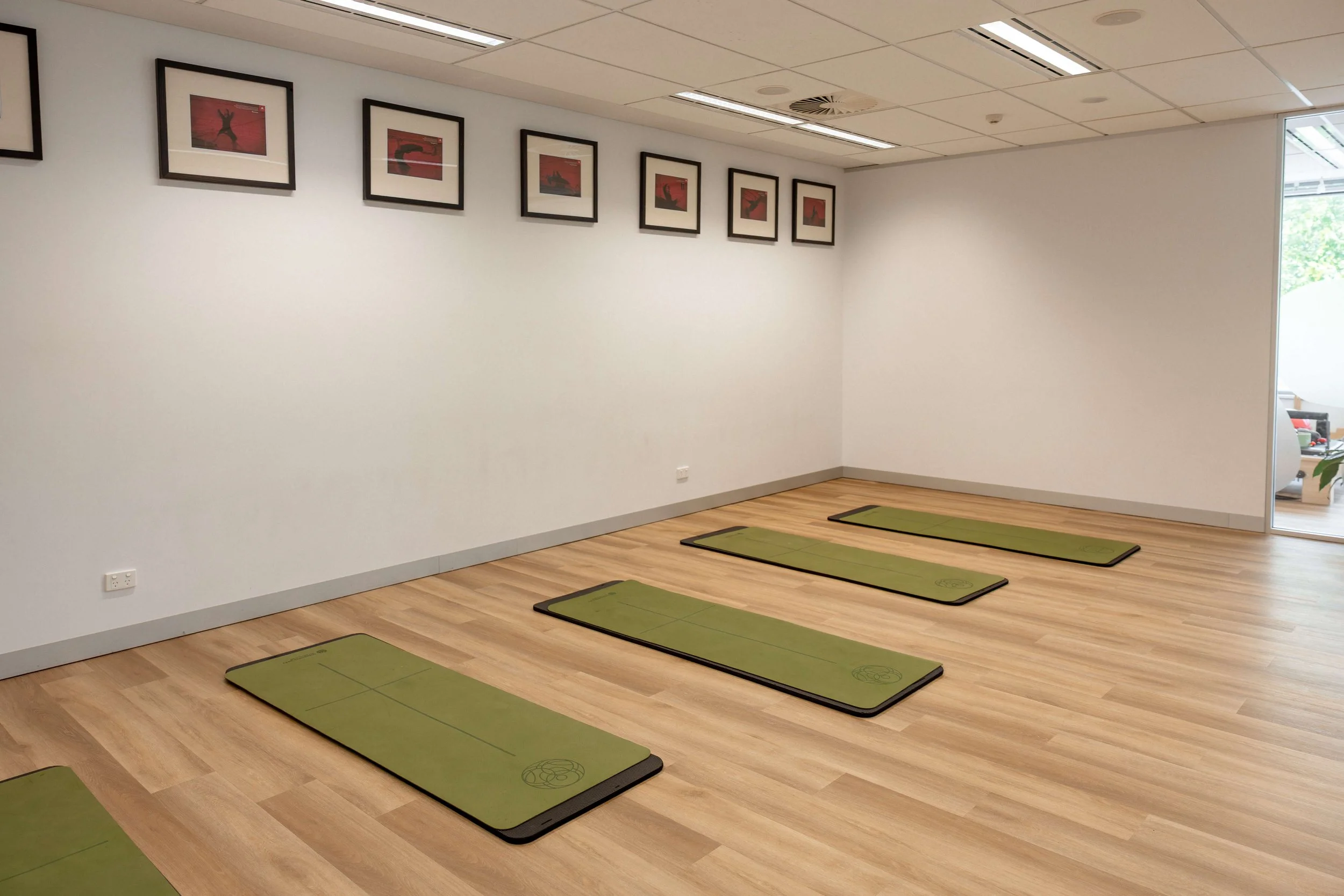 An empty fitness or yoga studio with six green yoga mats evenly spaced on a wooden floor, white walls decorated with framed abstract artwork, and a glass door leading outside.