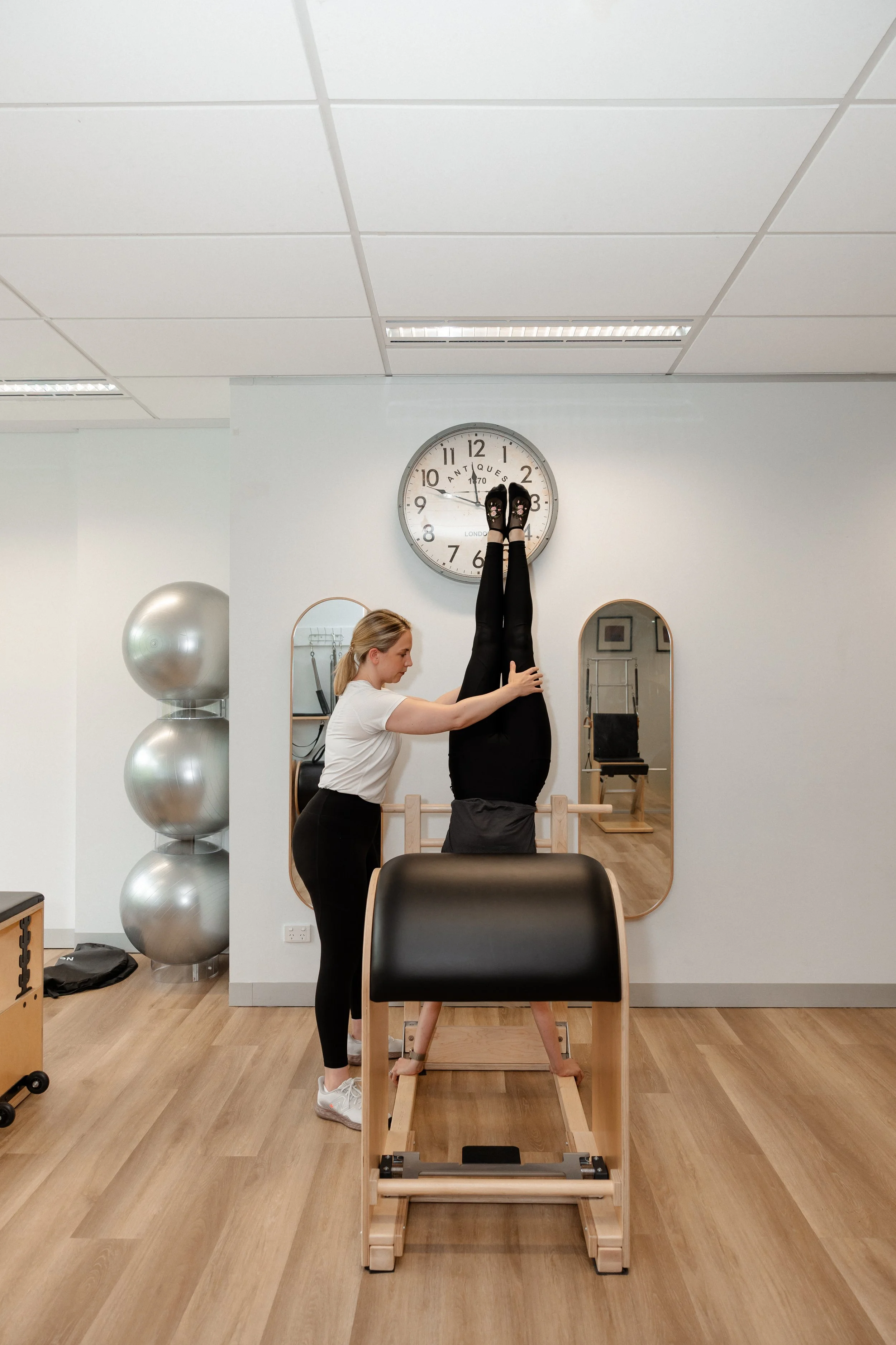 A woman assisting another person who is doing a handstand against a wall with feet touching a clock on the wall in a fitness studio.