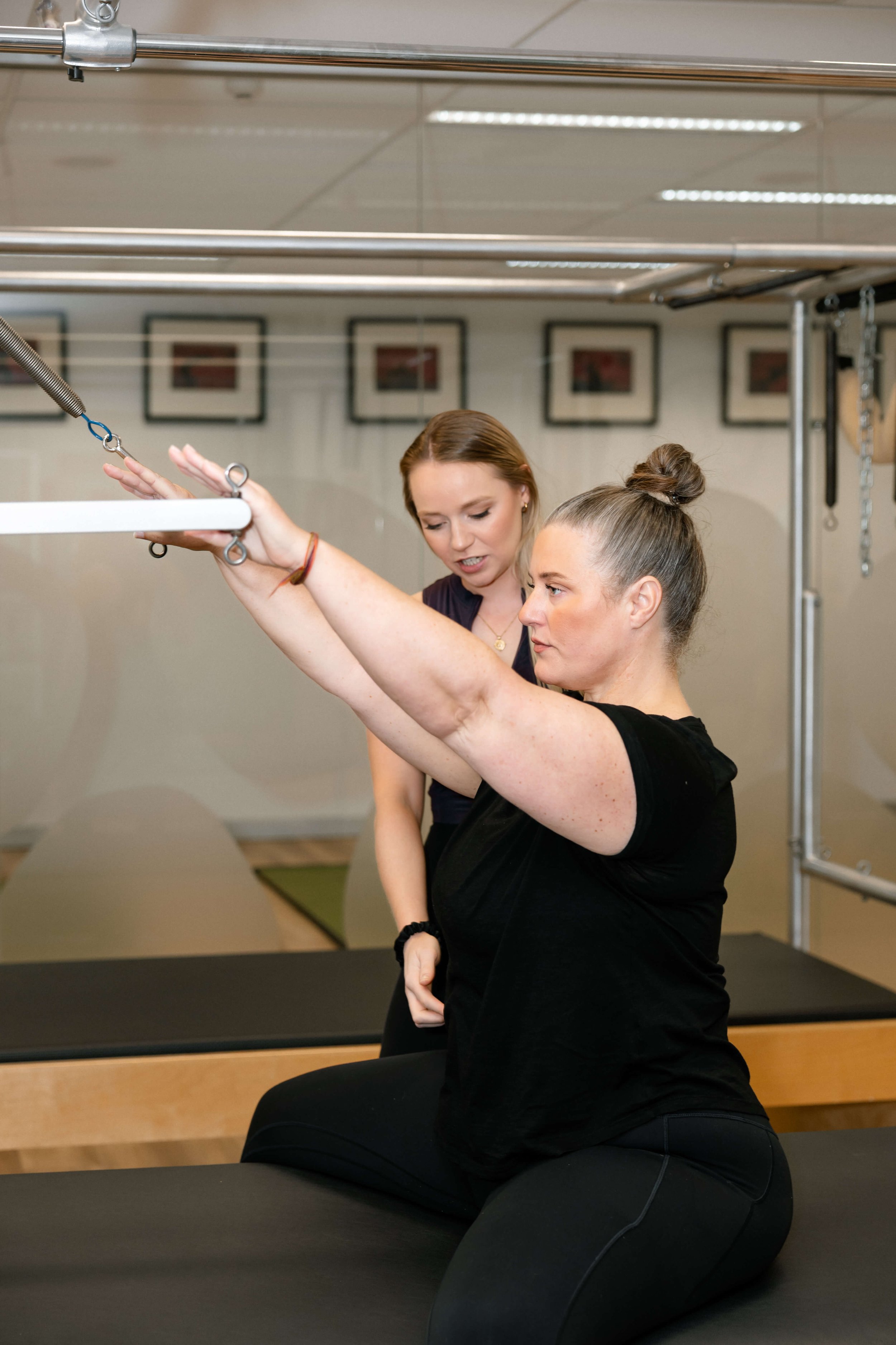 A woman is performing a physical exercise on a Pilates reformer machine while a trainer guides her. They are in a fitness studio with framed pictures on the wall.