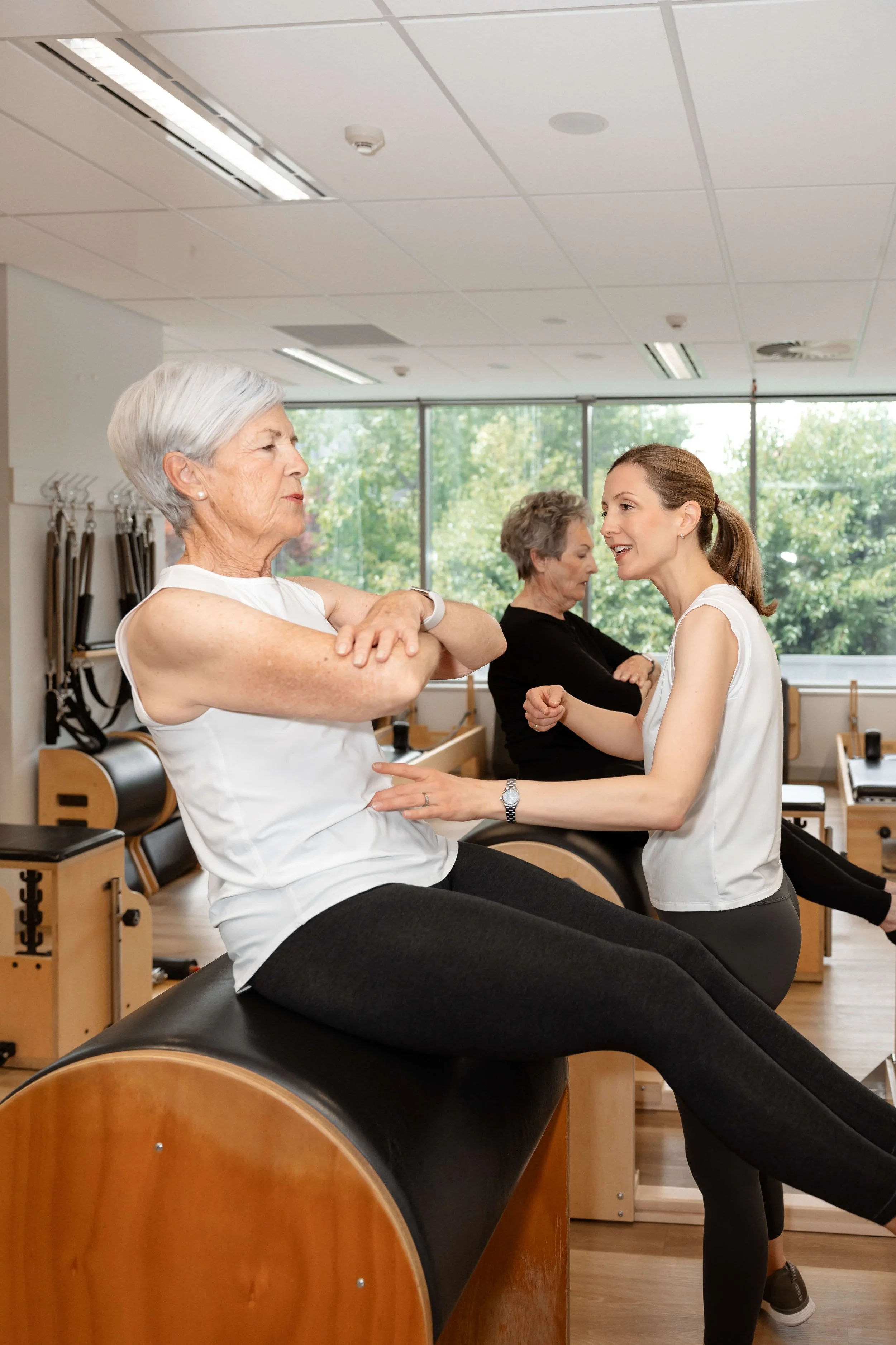 An older woman exercising on a Pilates reformer with a trainer holding her hands, in a fitness studio with large windows and other women exercising in the background.