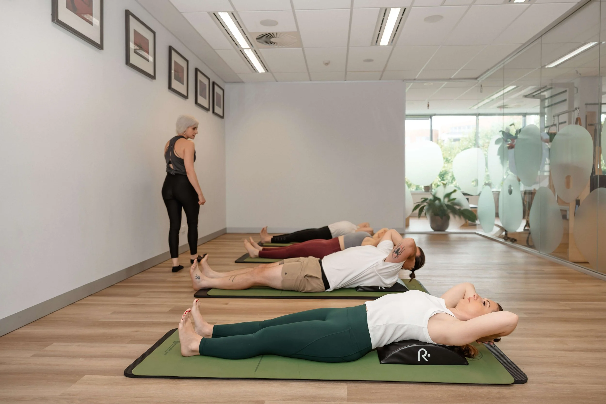 A yoga class with five women lying on mats in a room with large windows, a woman standing nearby observing or instructing, and art frames on the wall.