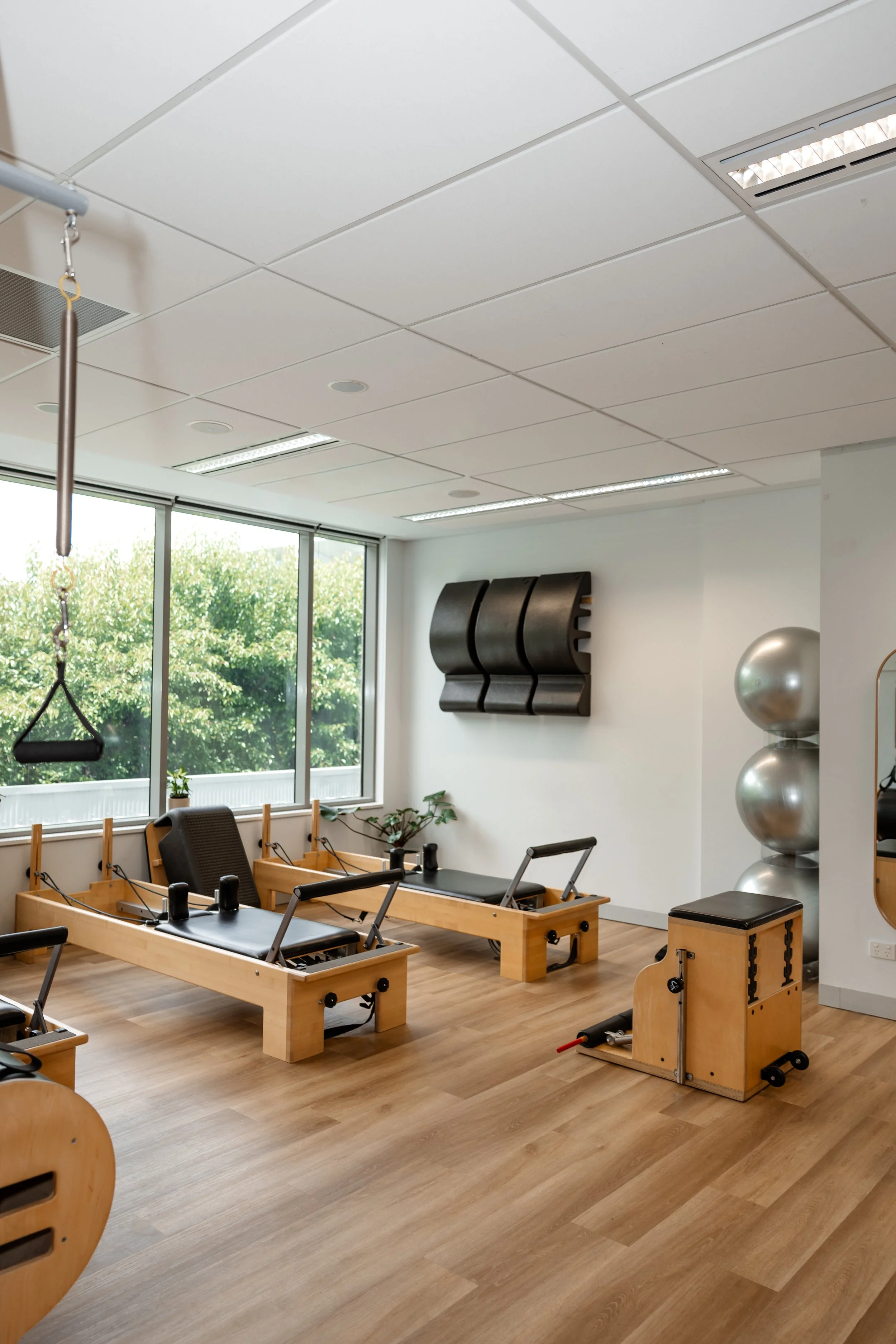  Pilates reformer machines in a well-lit fitness studio with large windows, exercise balls, and wooden flooring.