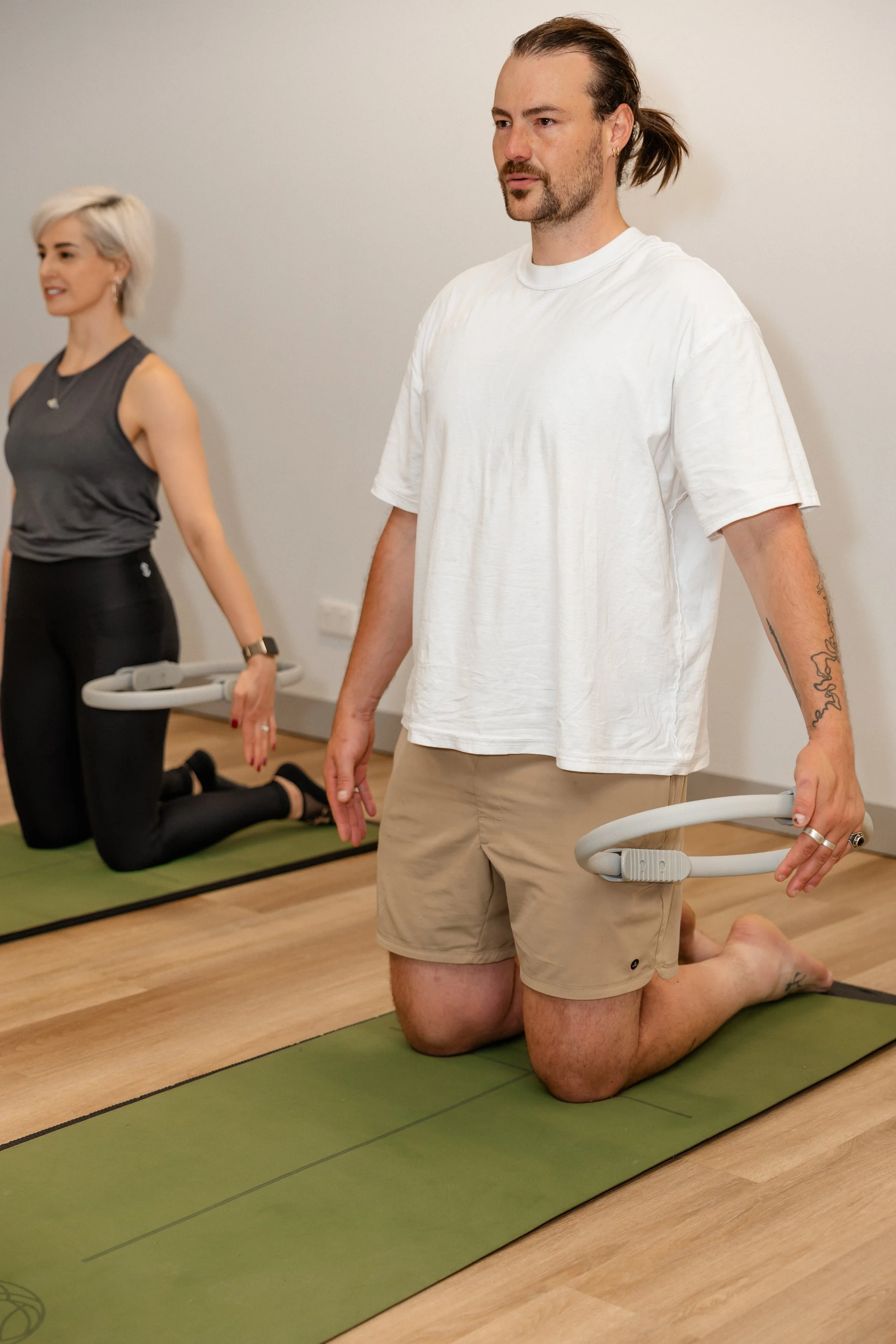 Two people practicing yoga, with the man in the foreground kneeling on a yoga mat holding a Pilates ring, and a woman in the background kneeling on a yoga mat.