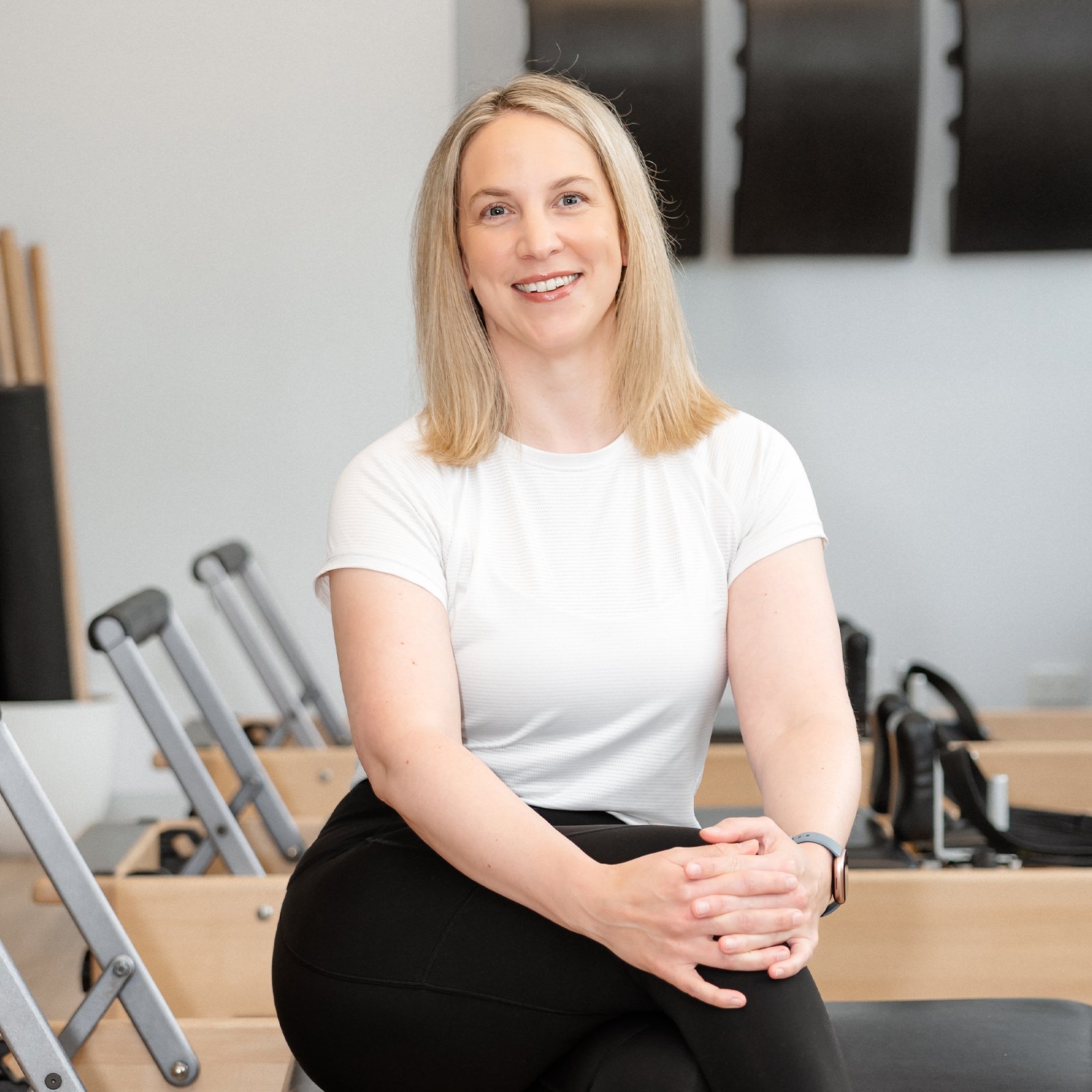 A smiling woman with shoulder-length blonde hair, wearing a white t-shirt and black pants, sitting on a Pilates reformer machine in a fitness studio.