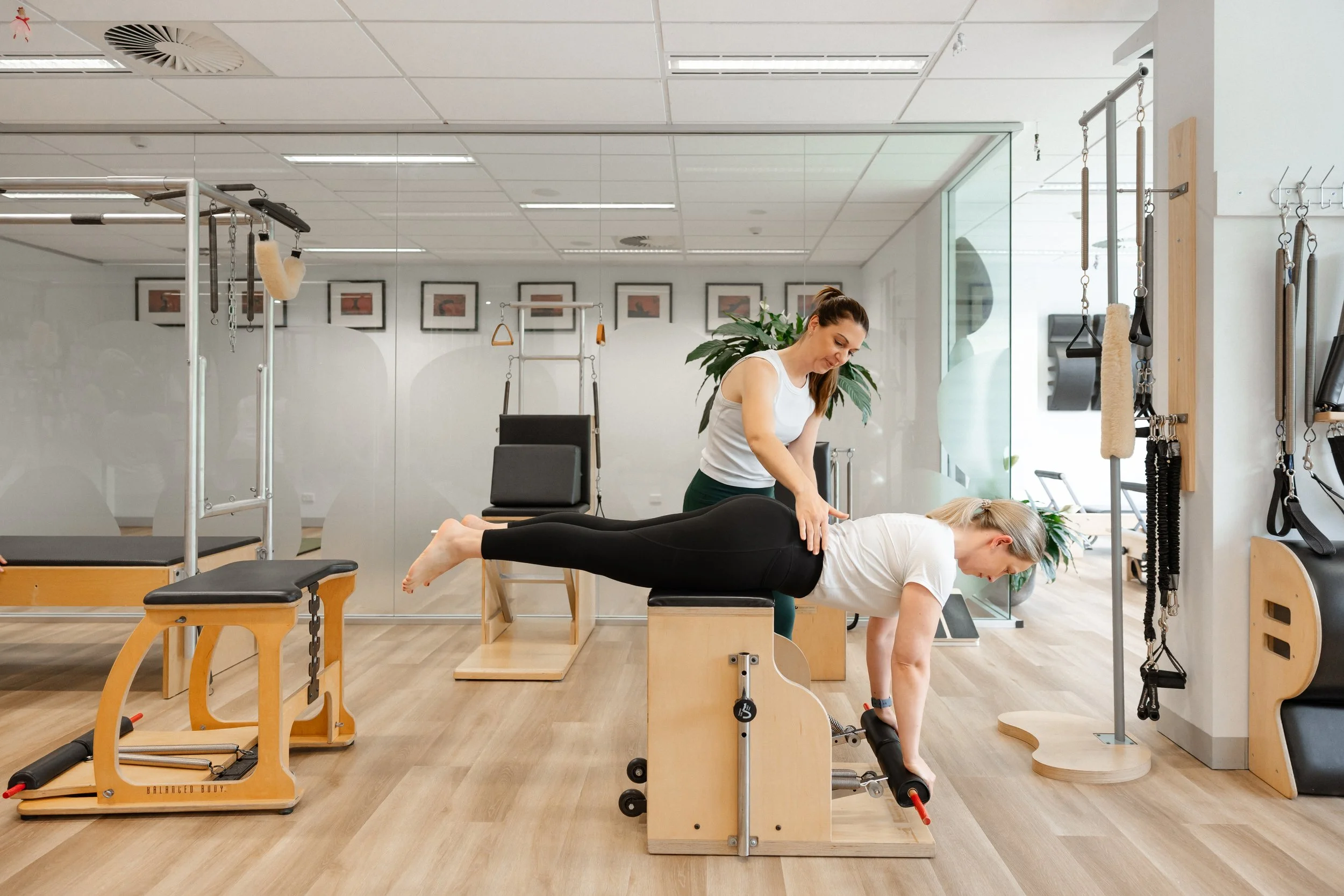 A woman practicing a plank exercise with her hands on a pilates reformer foot bar, while another woman assists her from behind in a fitness studio.