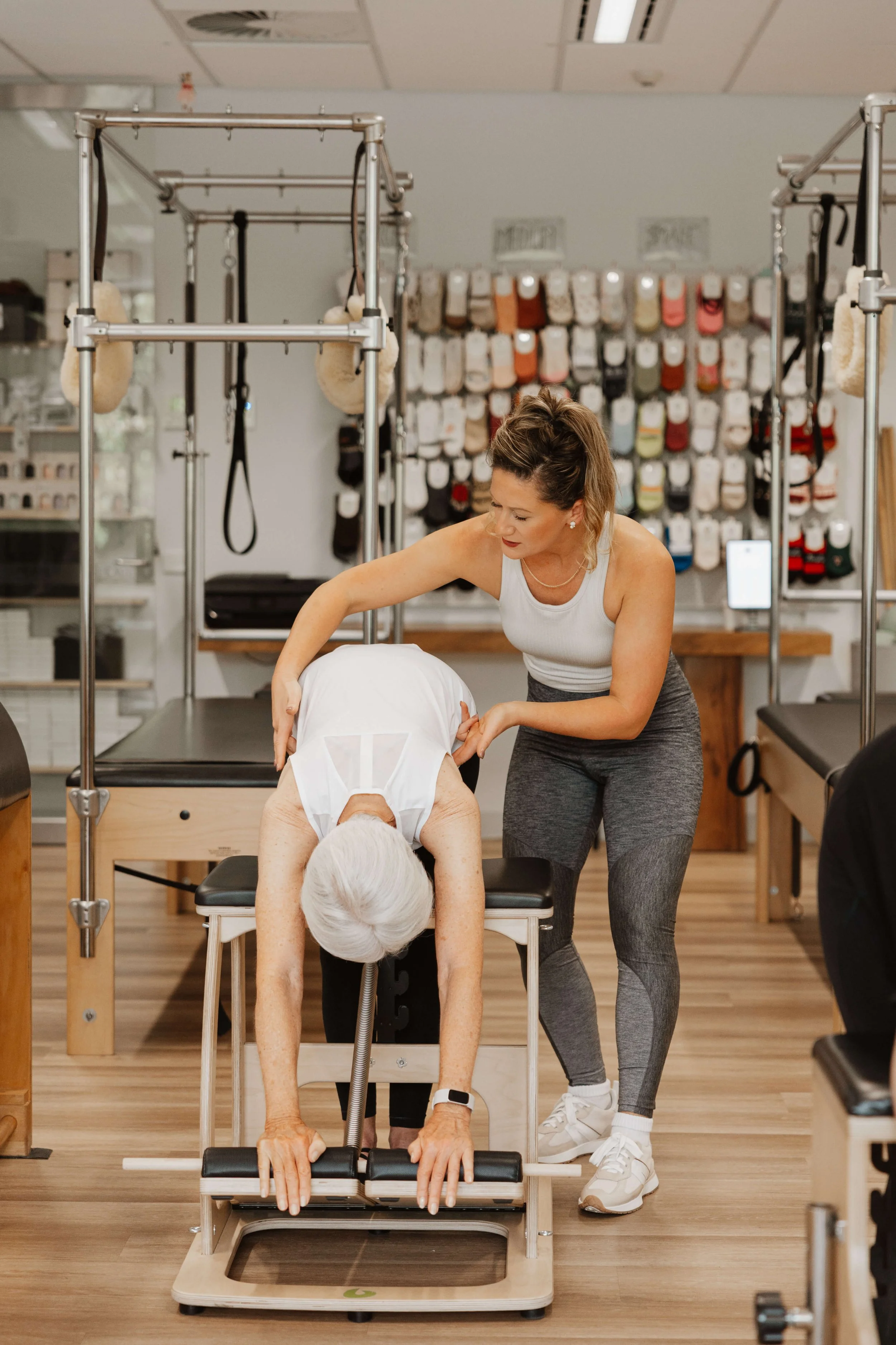 A physical therapist assisting an elderly woman with an exercise on a reformer machine in a therapy room. The room has mats, therapy equipment, and a wall of sock shelves in the background.