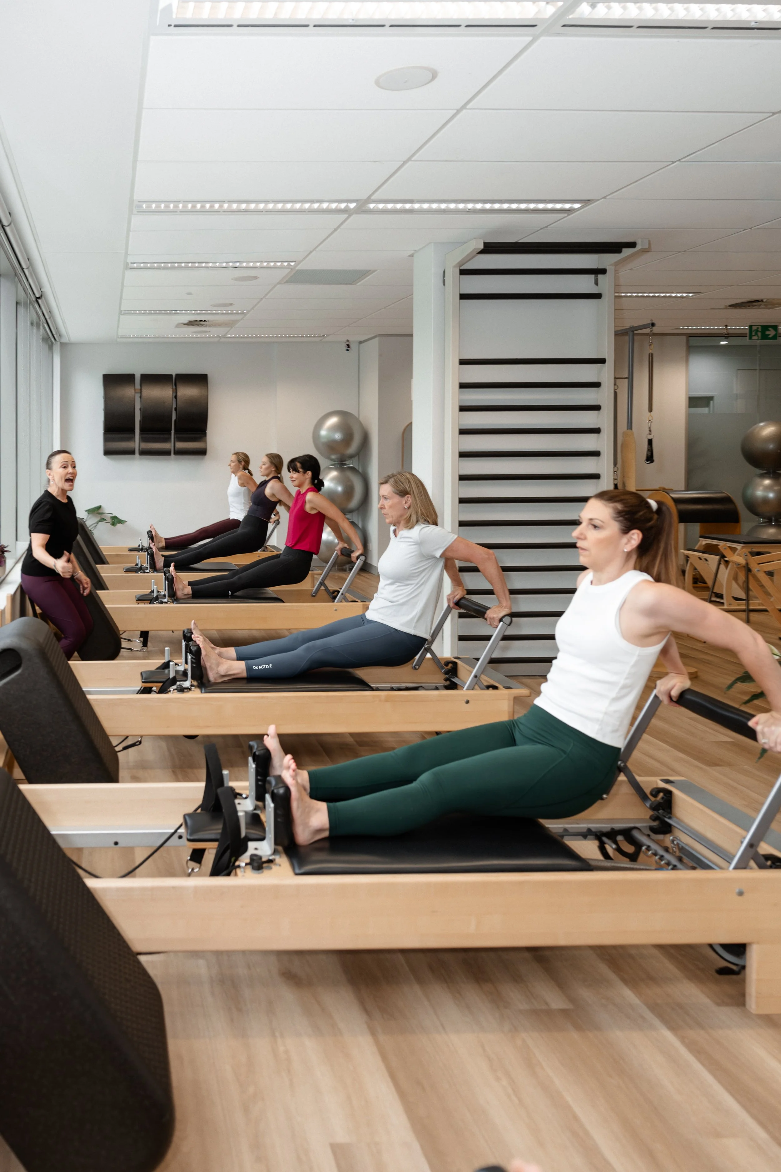 Women participating in a Pilates or fitness class on reformer machines in a fitness studio, with an instructor guiding them.