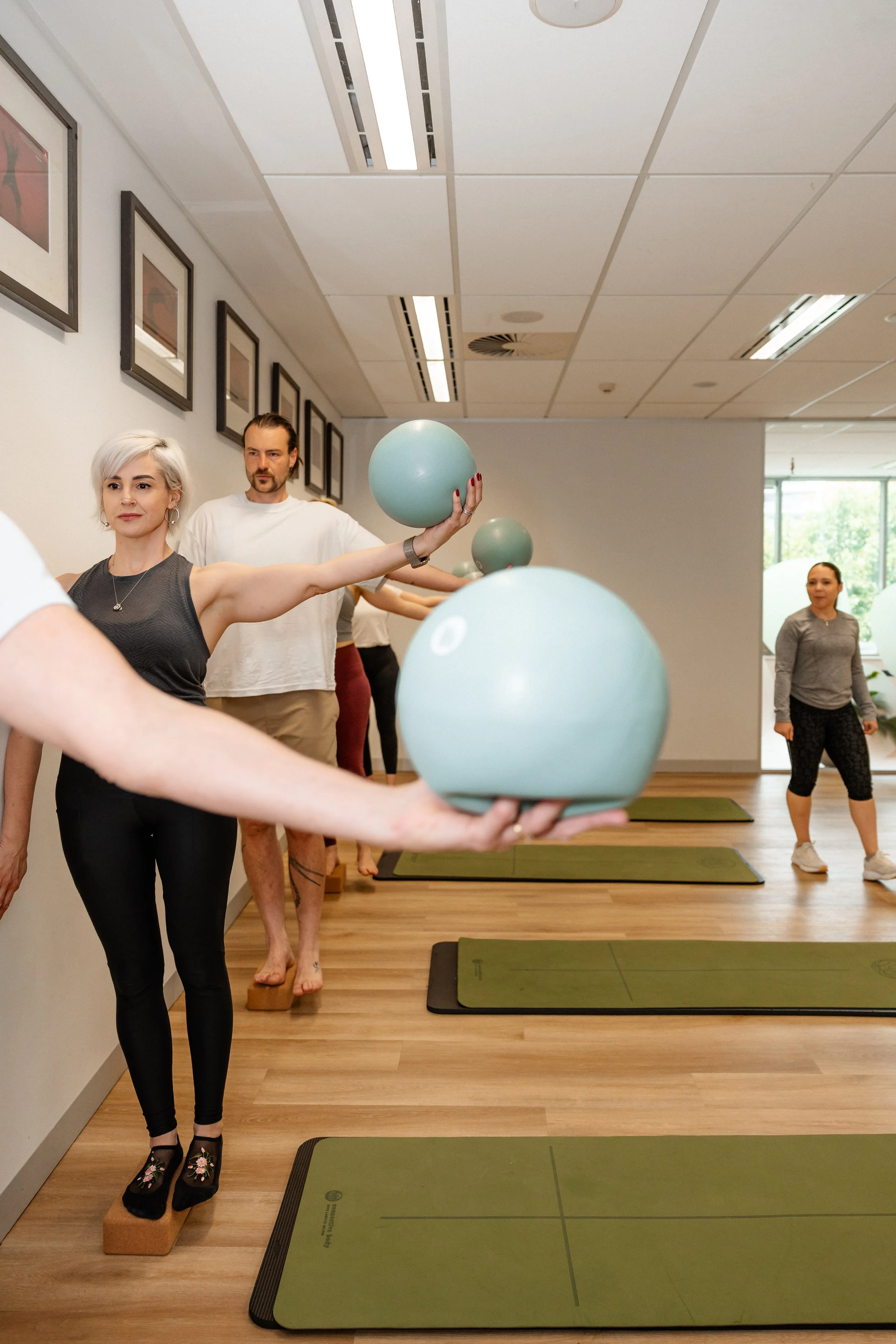 People participating in a yoga or meditation class, holding light blue round balls, standing on green mats, in a room with framed pictures on the wall and large windows.