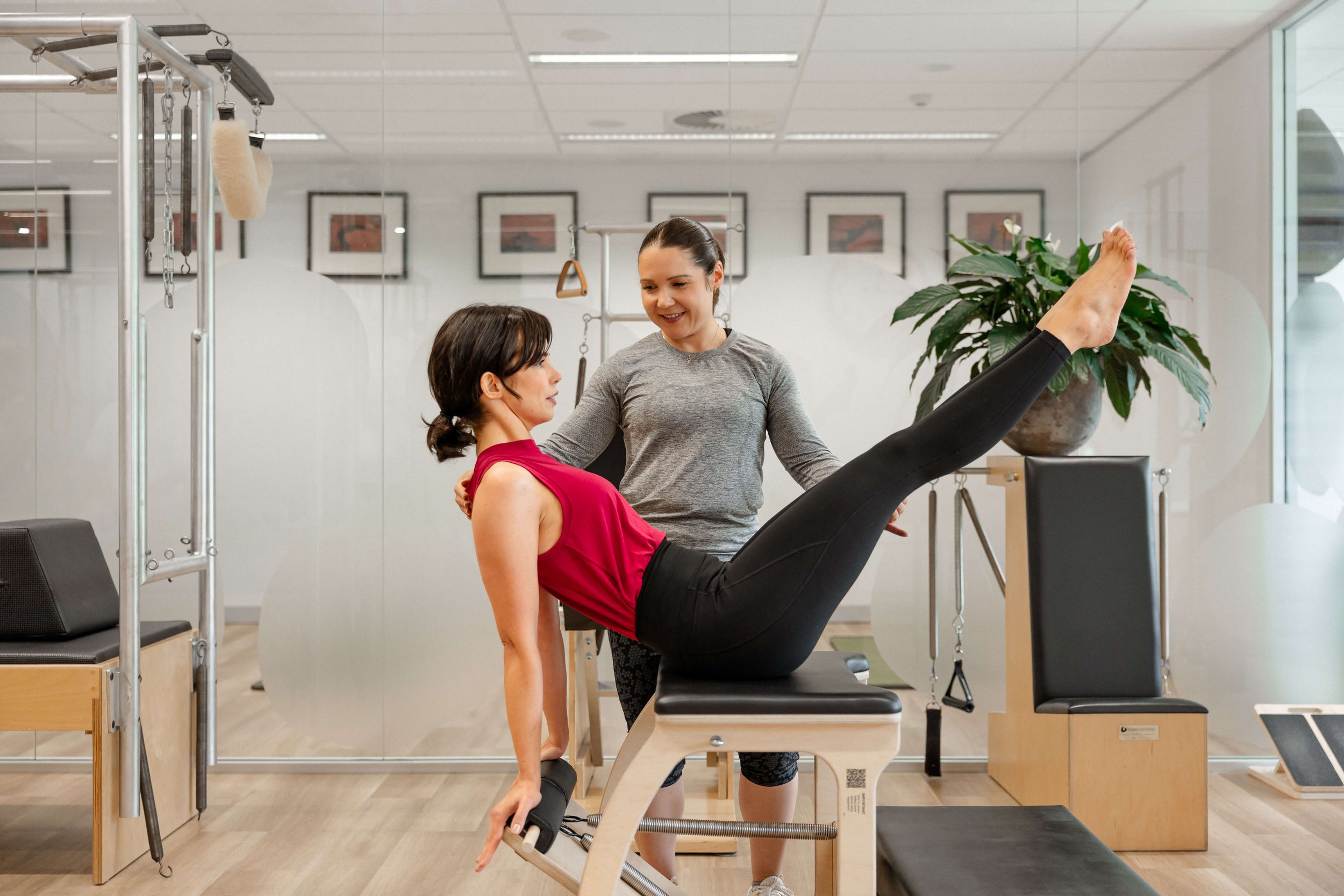 Physical therapist assisting woman in black leggings and a red tank top with leg exercises on a Pilates reformer machine in a well-lit physiotherapy clinic.
