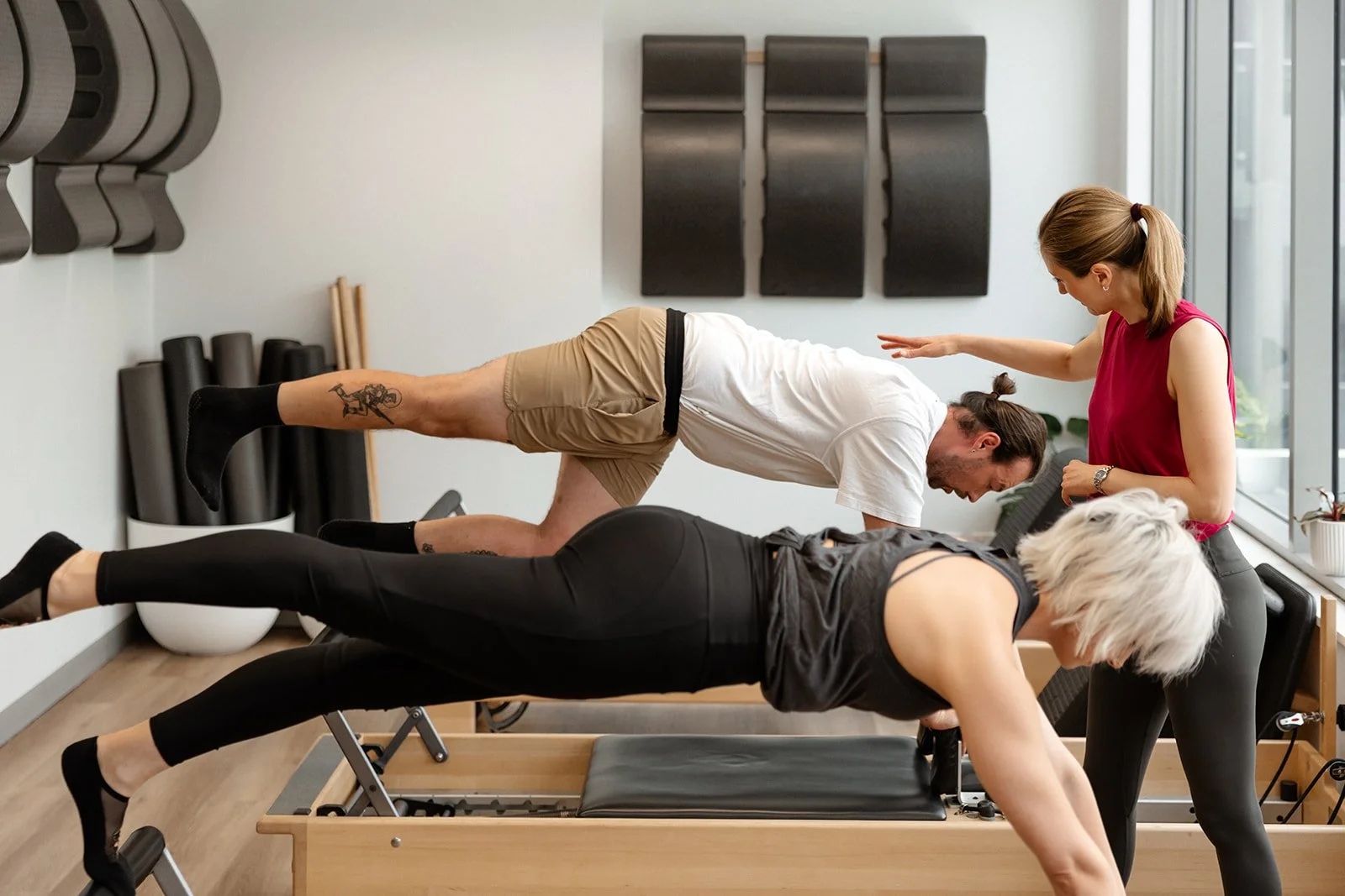 Two women and one man exercising on Pilates reformer machines in a fitness studio, with a trainer instructing them.
