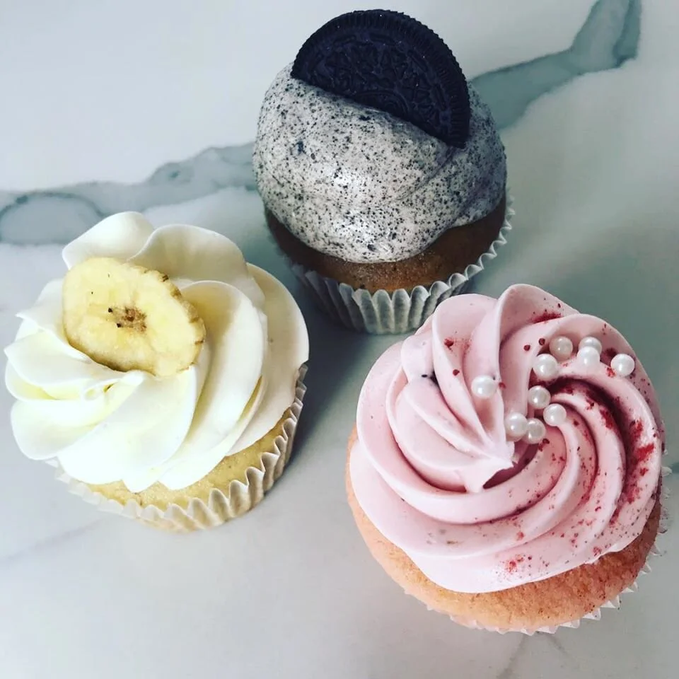 Three cupcakes on a white surface: one with white frosting and a banana slice, one with pink frosting and pearl sprinkles, and one with cookies and cream frosting with an Oreo cookie on top.