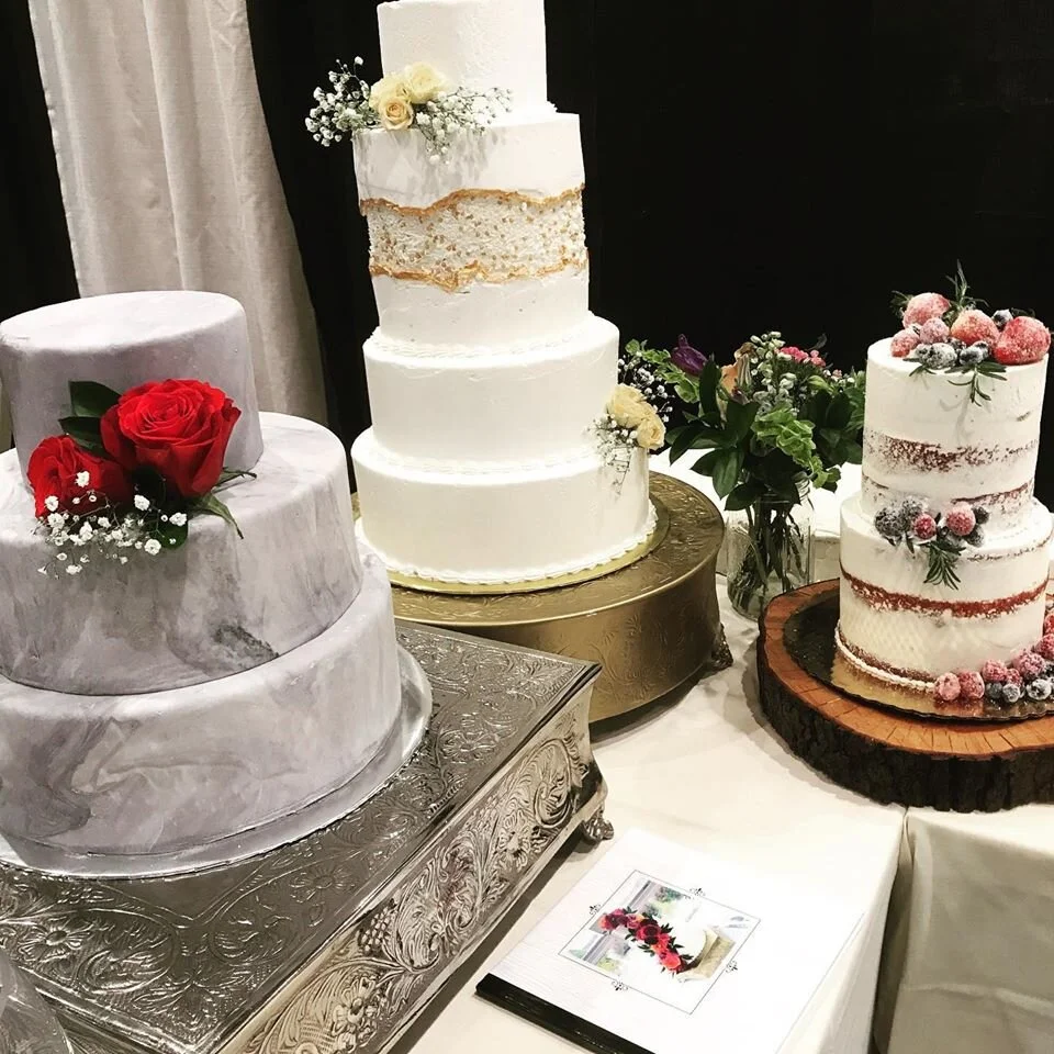 A display of three decorated wedding cakes on a table. The left cake is marble-patterned with red roses, the middle cake is a four-tier white cake with gold accents and flowers, and the right cake is a three-tier semi-naked cake with berries and flowers on a wooden slab.