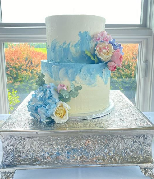 Two-tier white and blue watercolor cake with pastel pink, purple, and yellow flowers on top, placed on an ornate silver cake stand near a window.