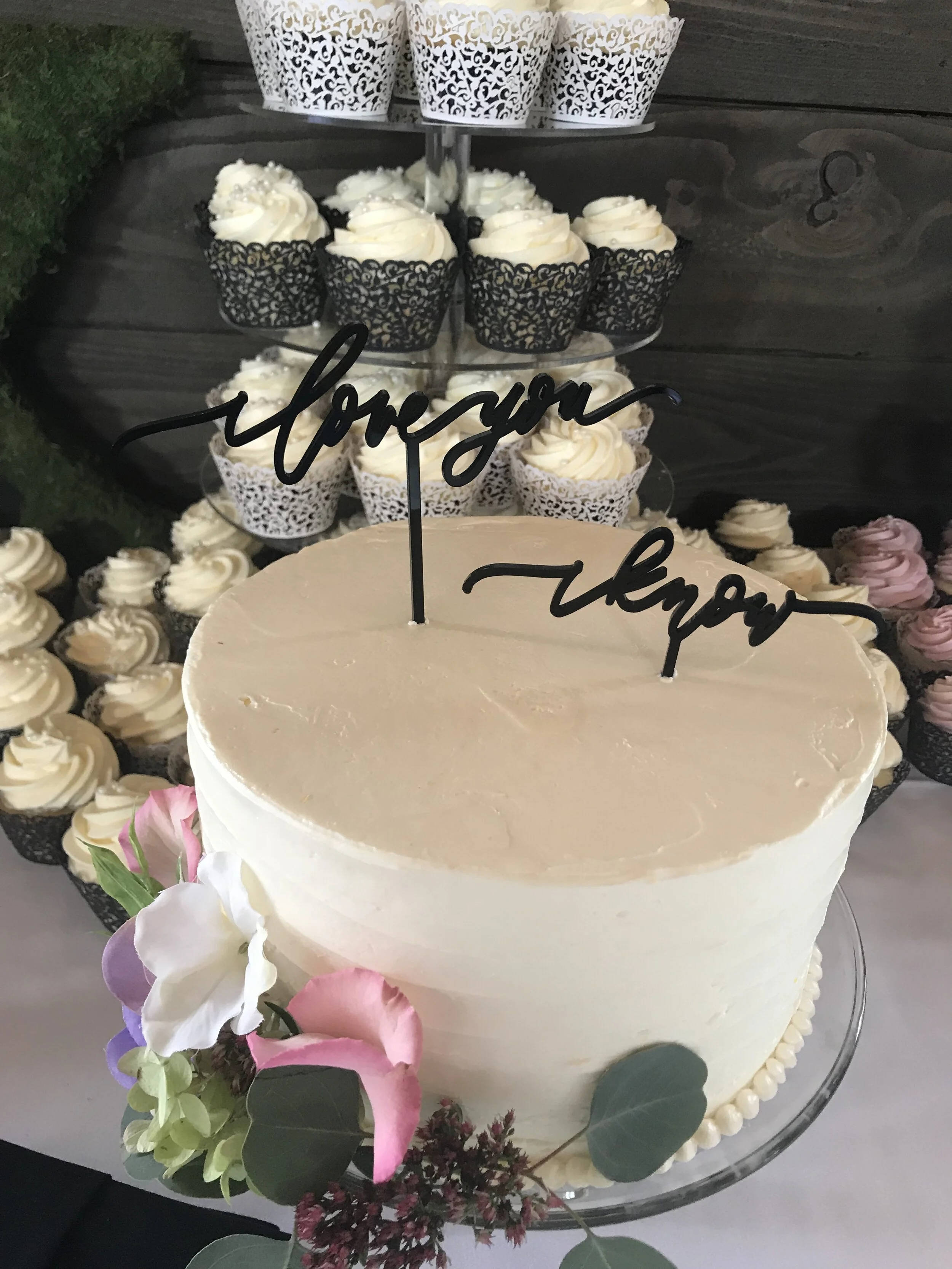 A wedding cake decorated with flowers and greenery, topped with black script cake toppers that say 'love you' and 'never'. In the background, there are multiple cupcakes with white and purple frosting, some in decorative black and white wrappers.