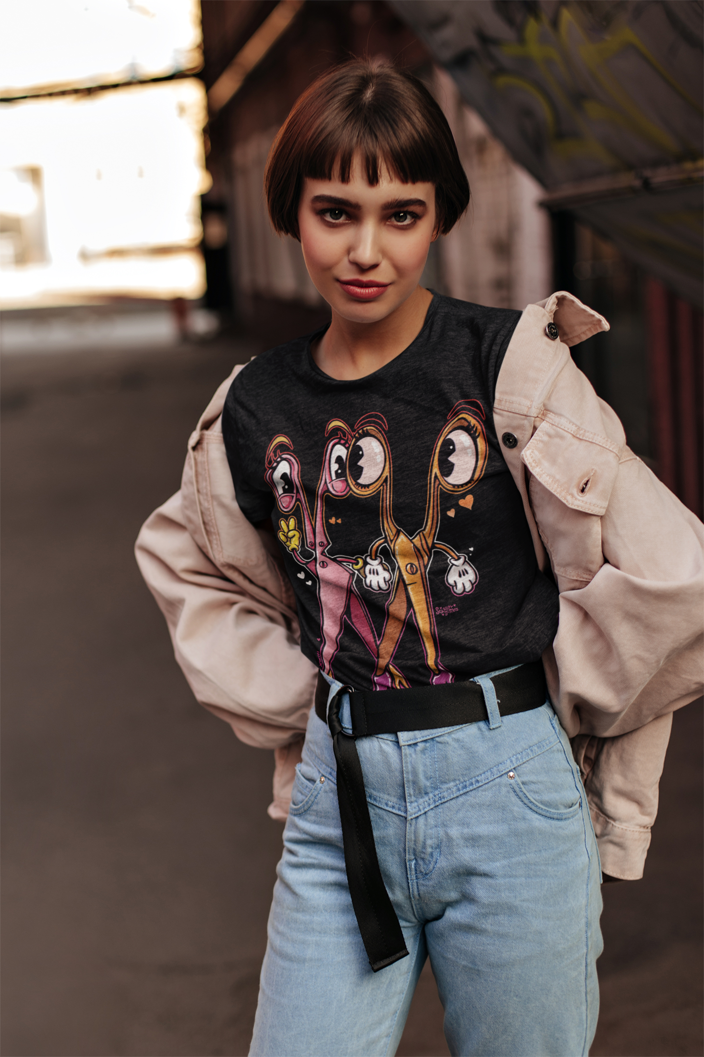 Joven con camiseta de personajes cartoon, chaqueta beige y pantalones vaqueros en un entorno urbano.