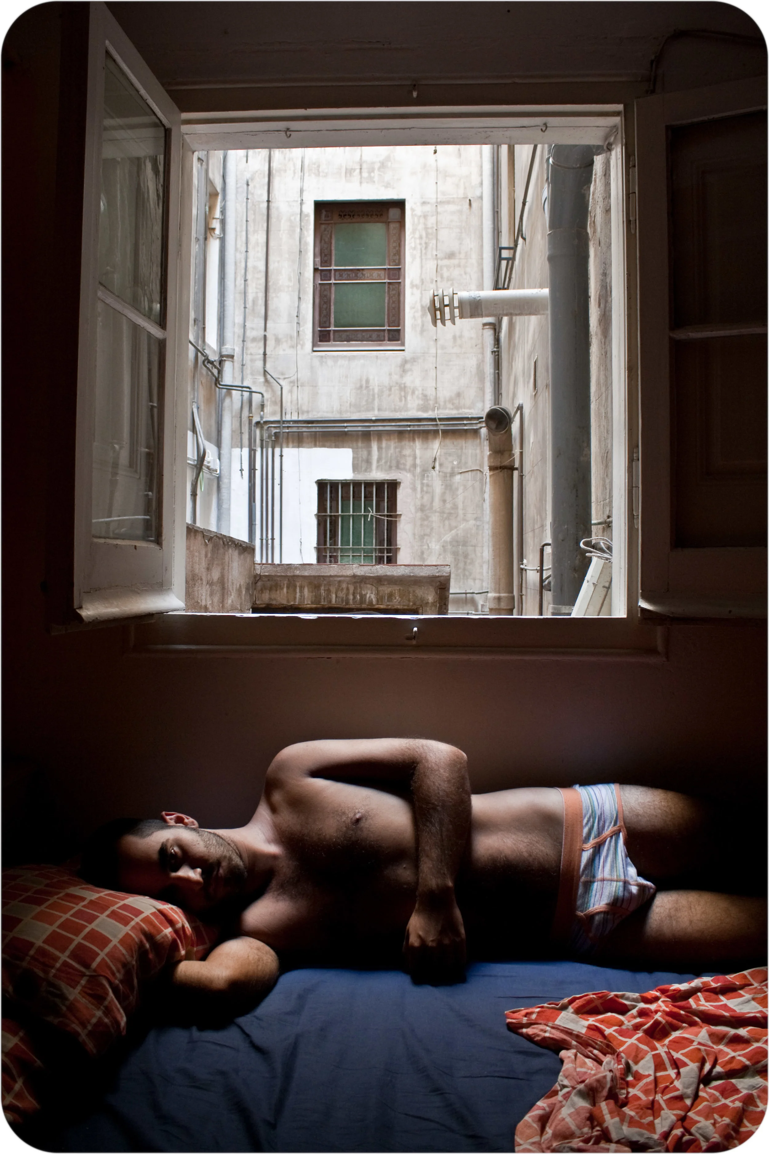 A shirtless man lying on a bed in front of an open window, with an urban view of buildings and pipes outside.