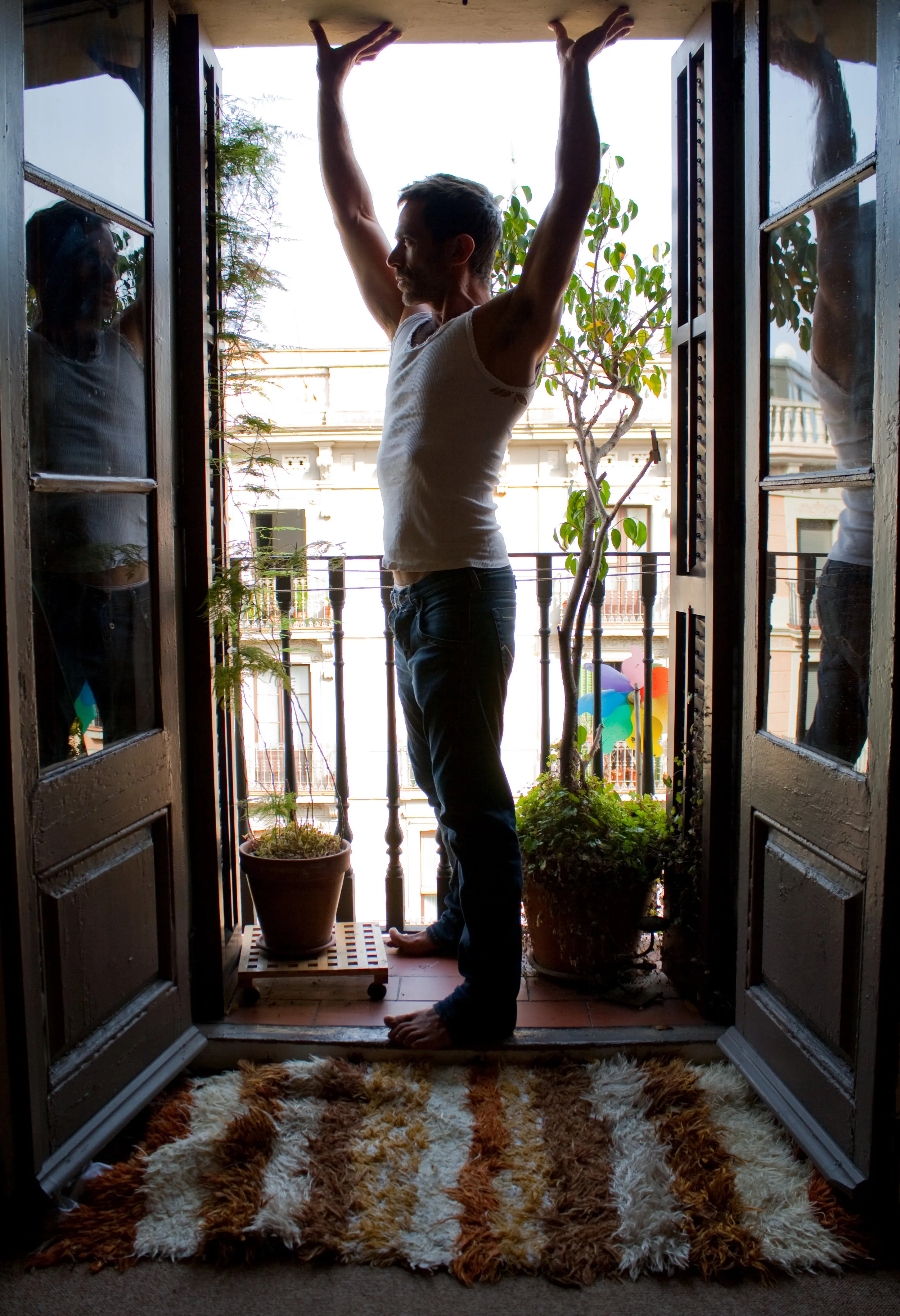 A man standing on a balcony, stretching with arms raised, seen through open balcony doors with reflections on the glass. There are potted plants and a colorful wind spinner outside, with sunlight illuminating the scene.