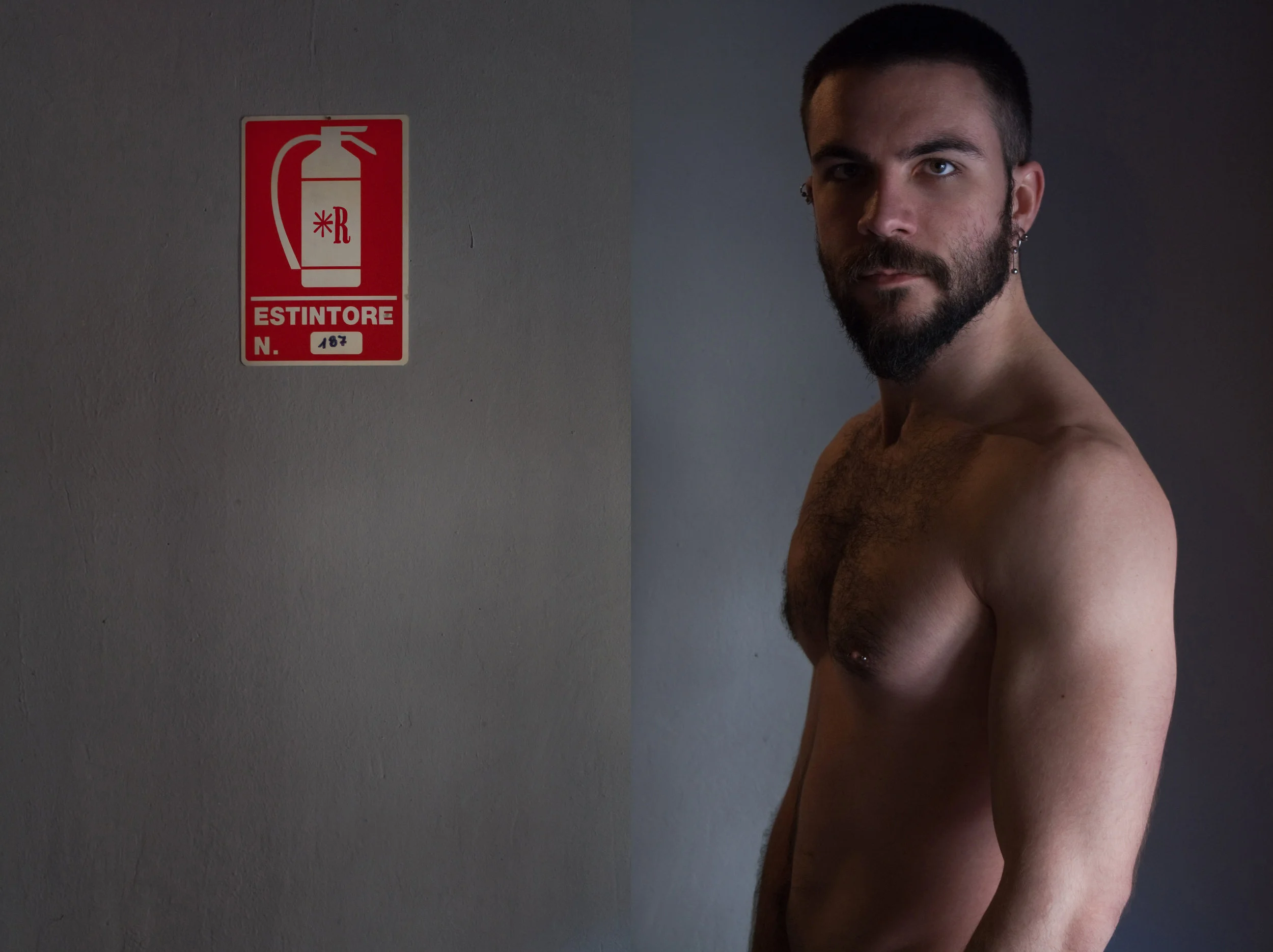 A shirtless man with a beard and earrings standing next to a wall with a fire extinguisher sign.