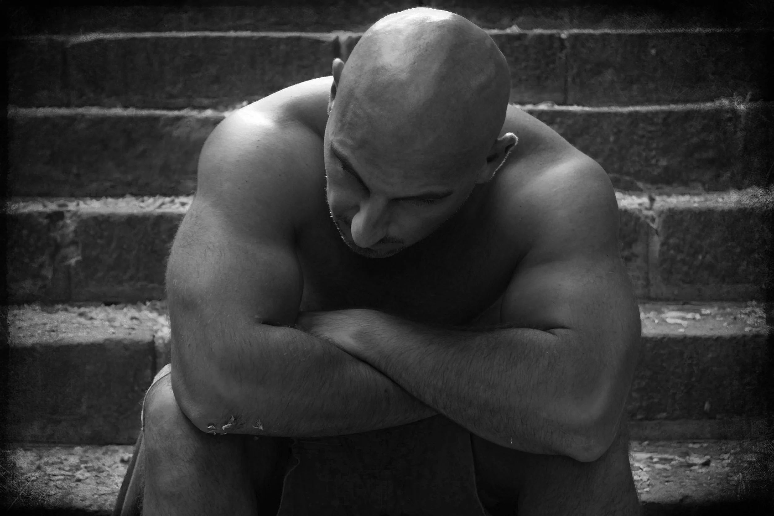 Black and white photo of a muscular, bald man sitting on stairs with his arms crossed and head bowed.