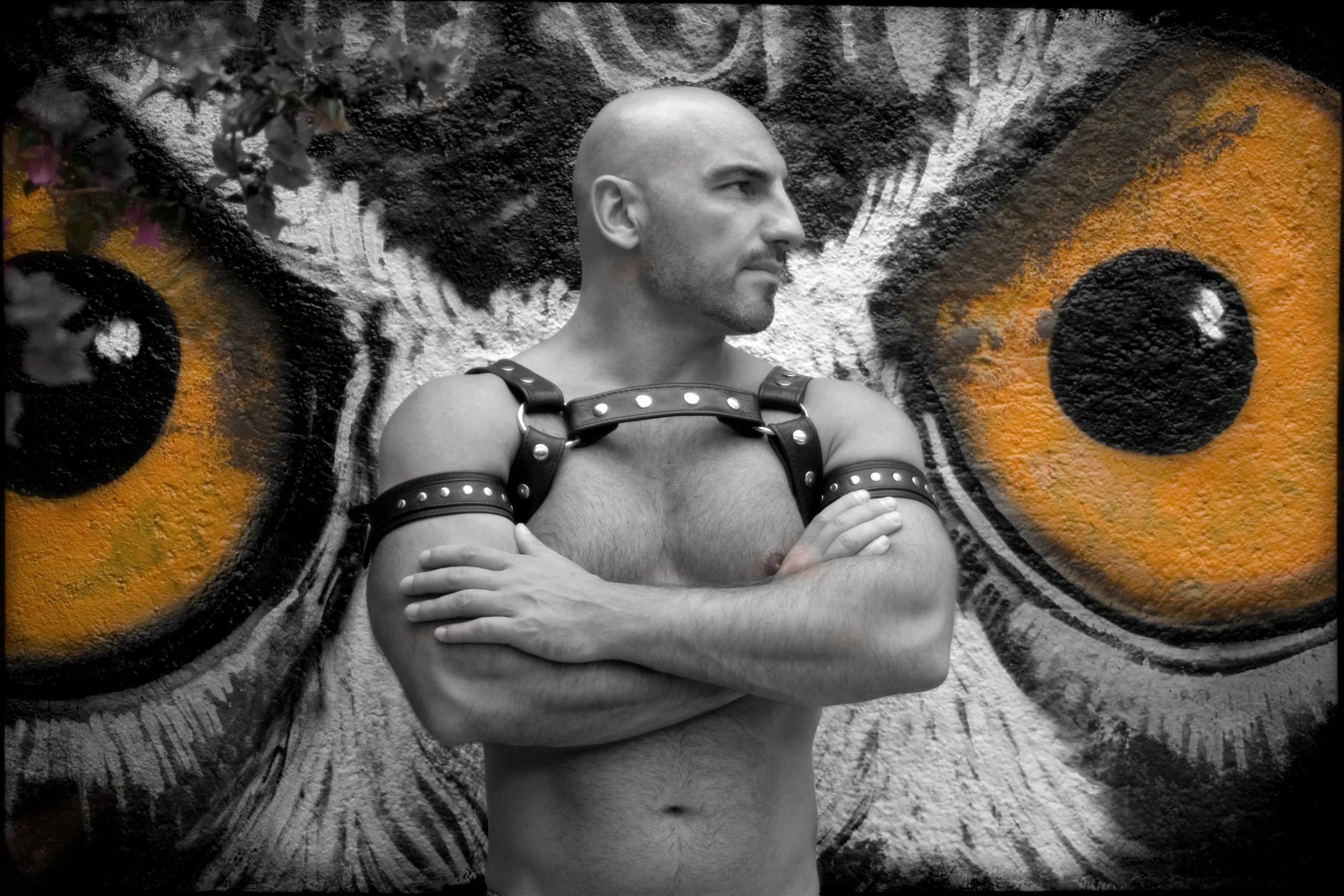A shirtless man with arms crossed in front of a graffiti mural of a butterfly with orange and black eyes on its wings.