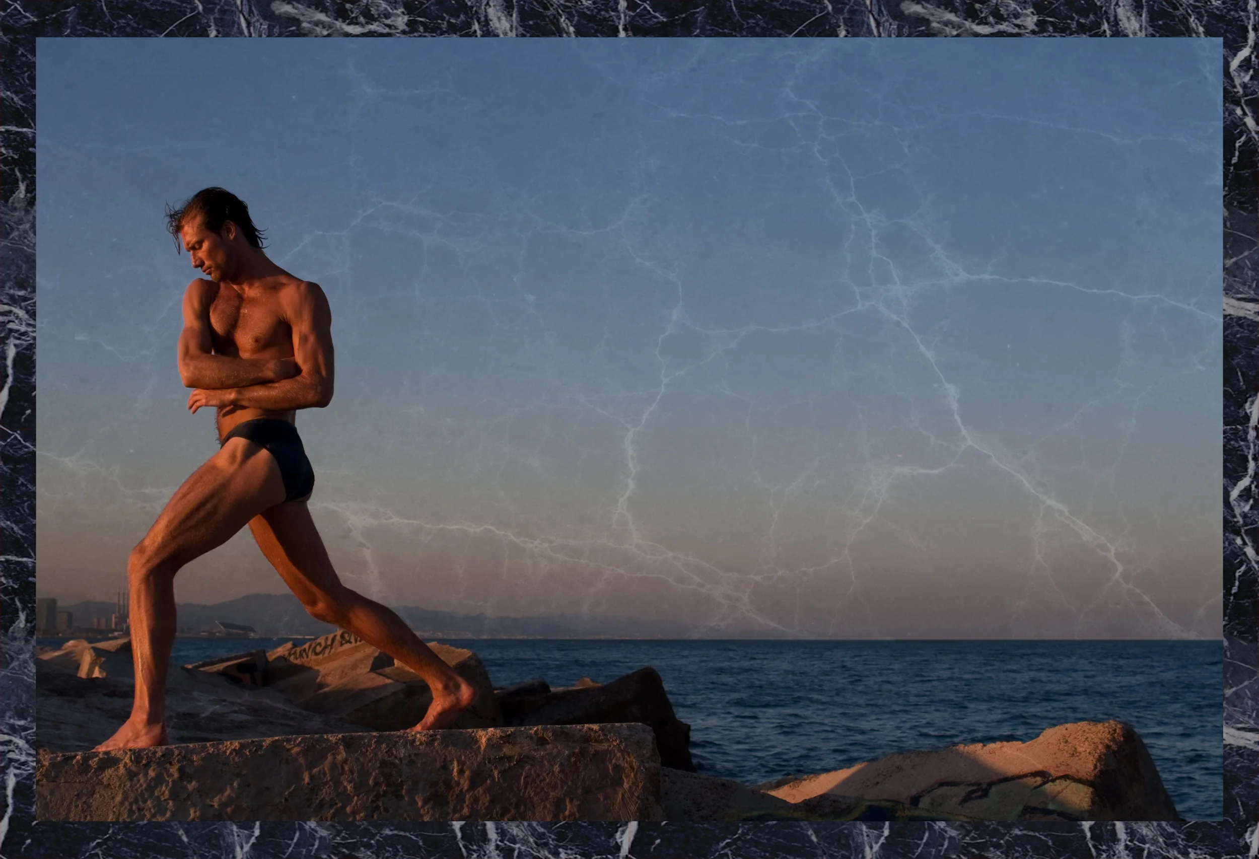 A man with a muscular build and long hair standing on rocks near the ocean during sunset. He is shirtless, wearing black shorts, with his arms crossed and looking down. The sky has a lightning pattern overlay effect, and the scene is framed with a bl