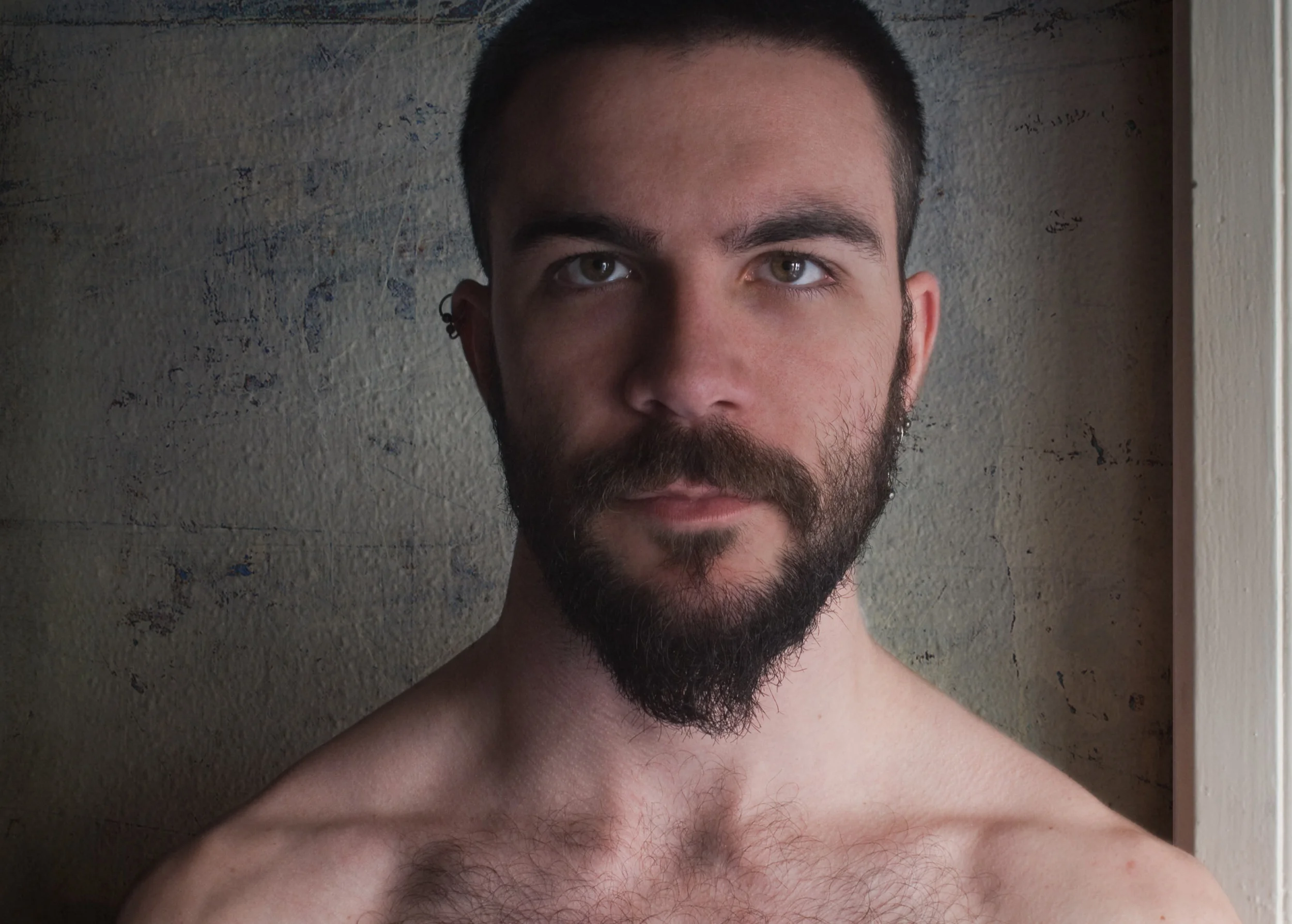 Close-up portrait of a shirtless young man with a beard and short dark hair, looking directly at the camera, standing against a textured wall.