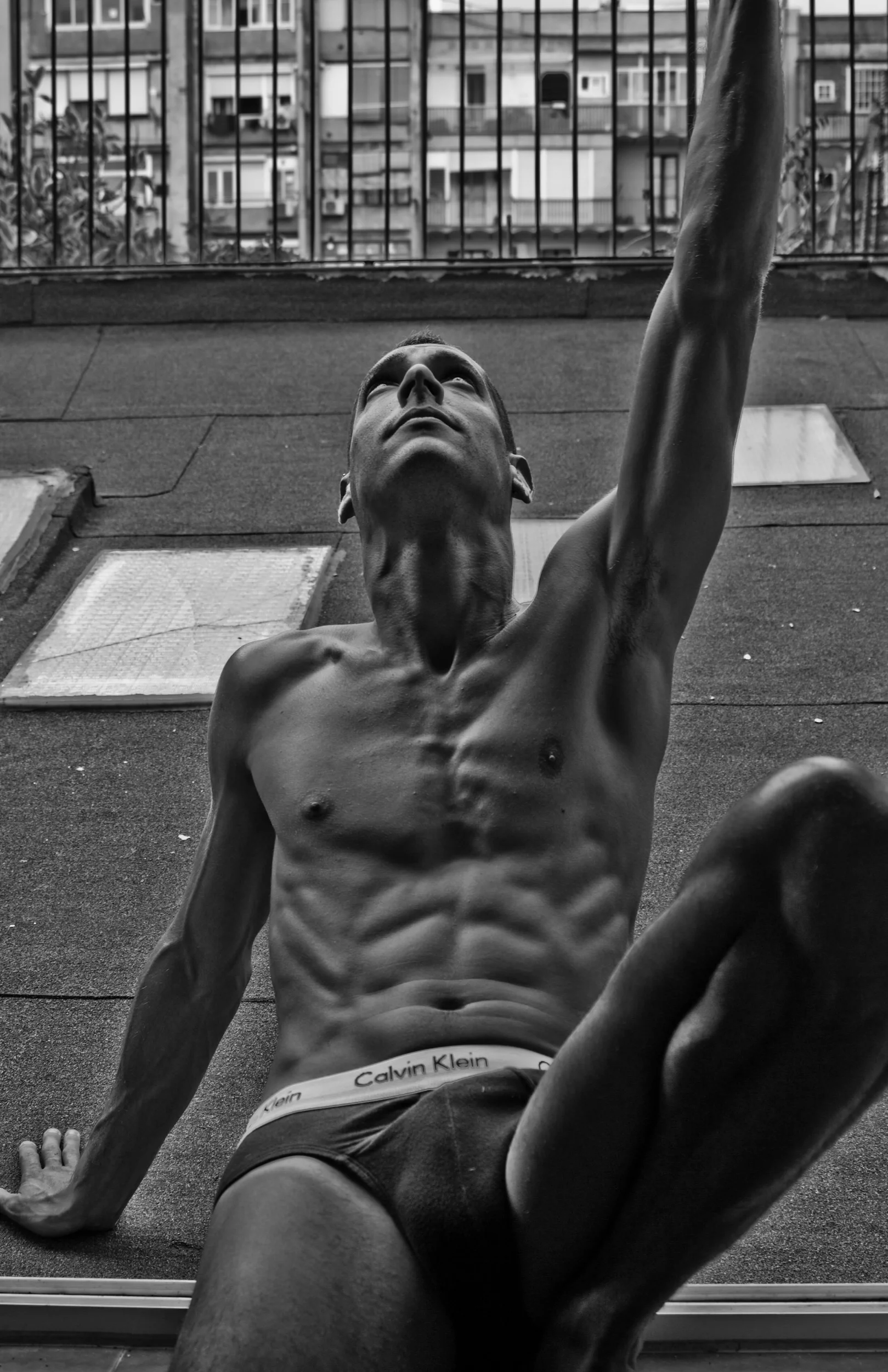 Black and white photo of a shirtless man with muscular abs, sitting on a rooftop ledge with a city skyline in the background, wearing Calvin Klein underwear.