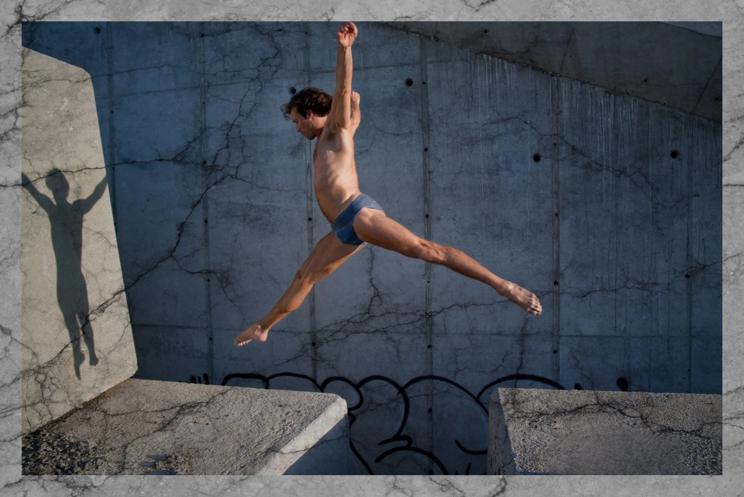 A man in gray shorts is mid-air performing a parkour jump between concrete structures, with his shadow visible on a concrete wall behind him.