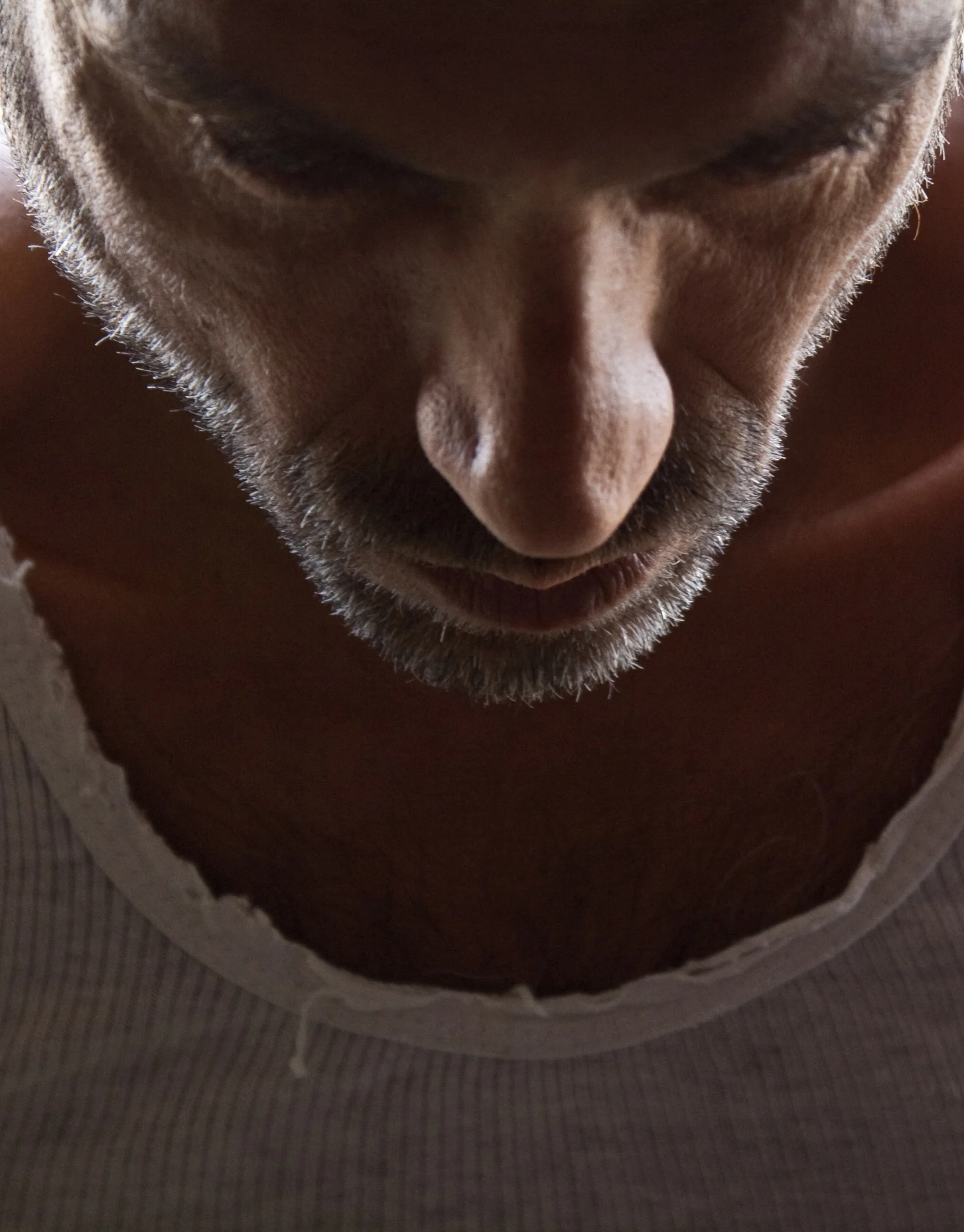 Close-up of a man's face looking downward, showing his nose, mouth, and part of his gray beard. He is shirtless and wearing a white tank top.