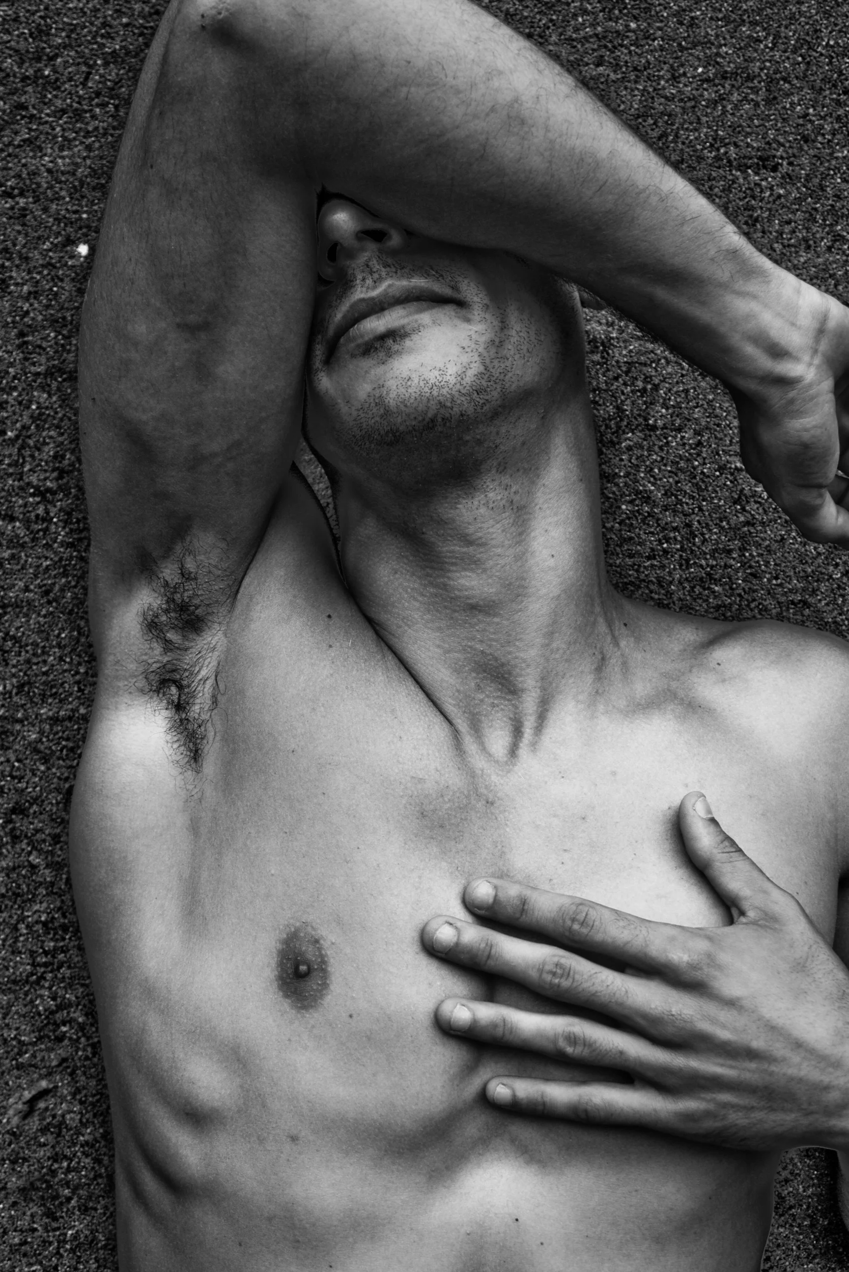 A black and white photograph of a shirtless man lying on the ground, with his arm covering his face and one hand touching his chest.