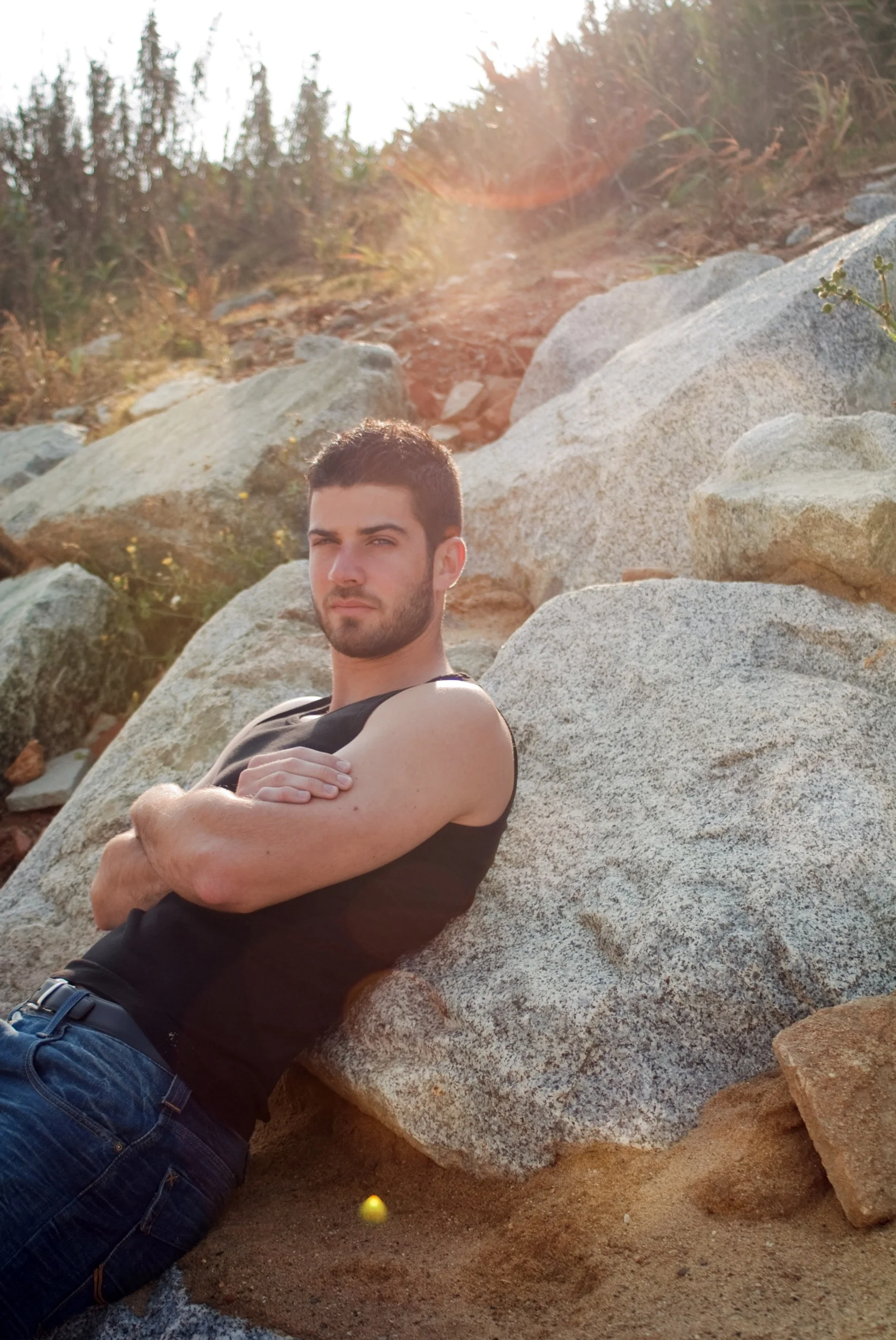 Young man with short dark hair and beard, wearing a black sleeveless shirt and jeans, leaning against a large rock outdoors with a forested area and sunlight in the background.