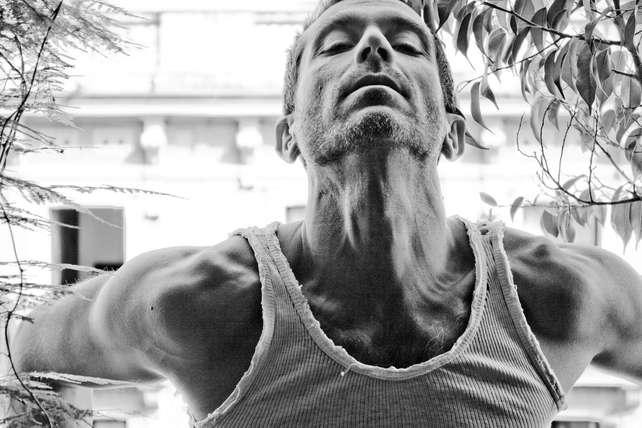 A close-up black and white portrait of a man with short hair and a beard, wearing a sleeveless shirt, with an upward-tilted head, surrounded by leaves.