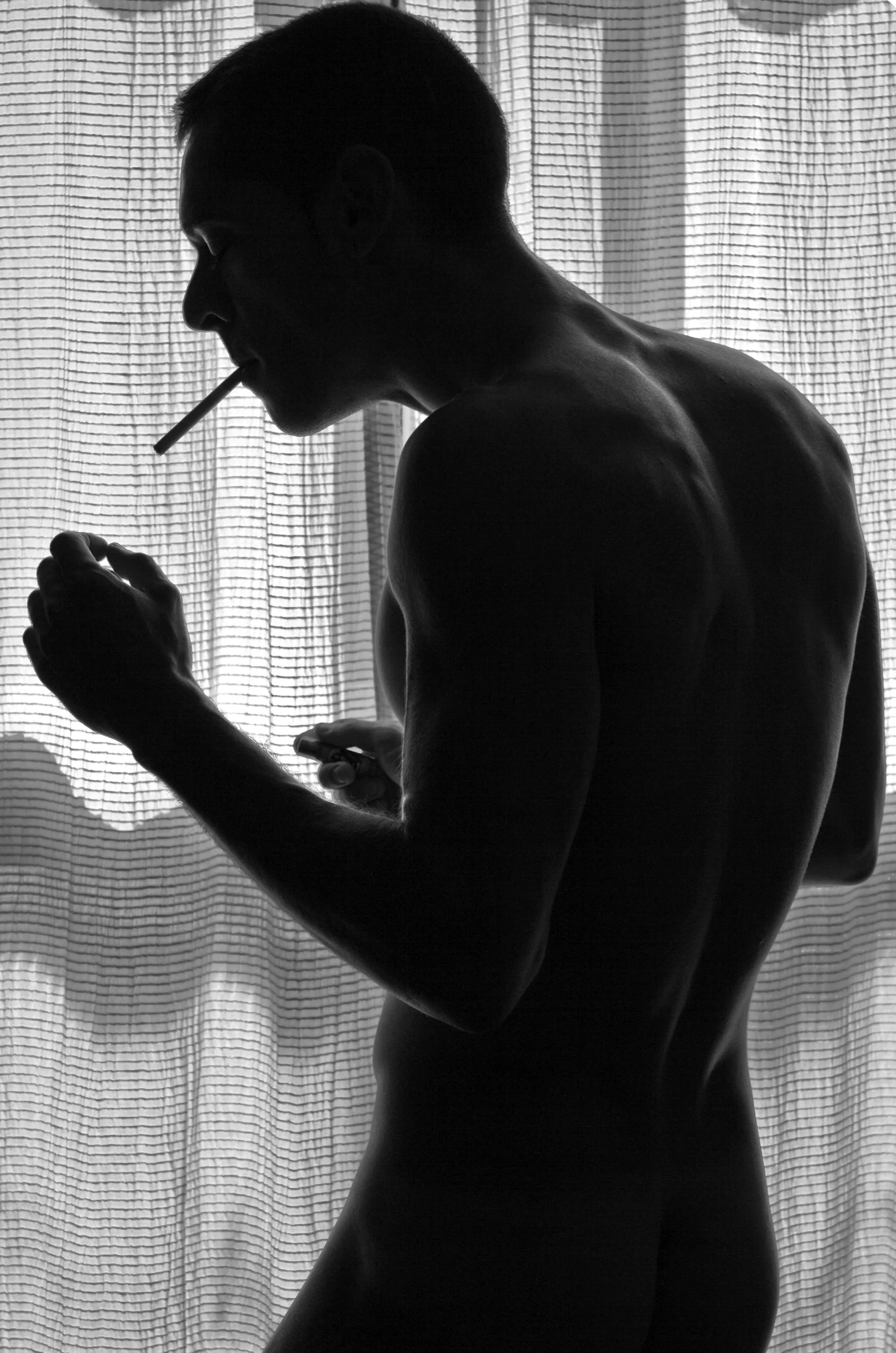 Silhouette of a shirtless man with a cigarette in his mouth, standing near a curtain, looking at his phone.