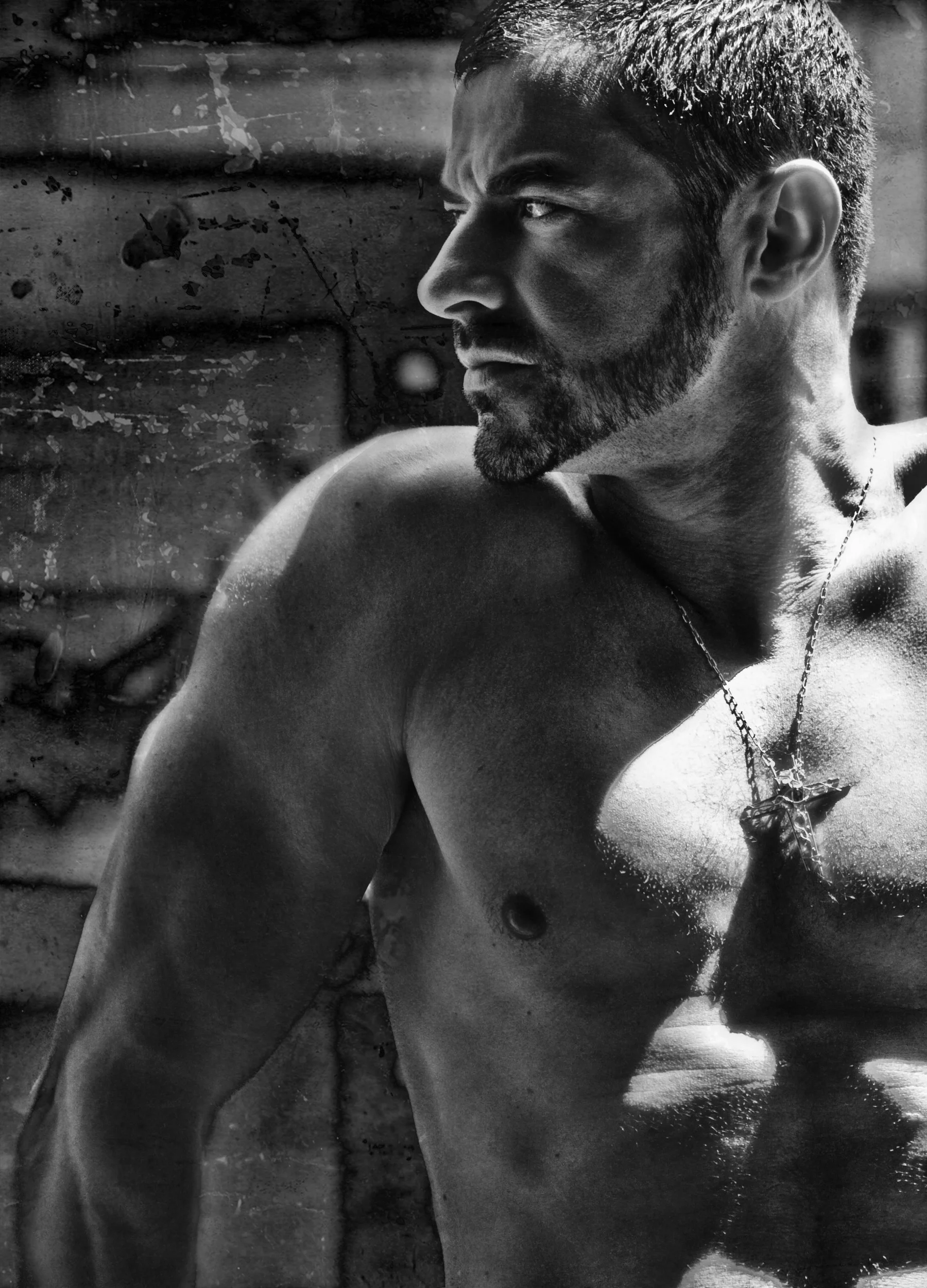 Black and white photo of a muscular man with a beard, looking to the right, wearing a necklace with a cross.