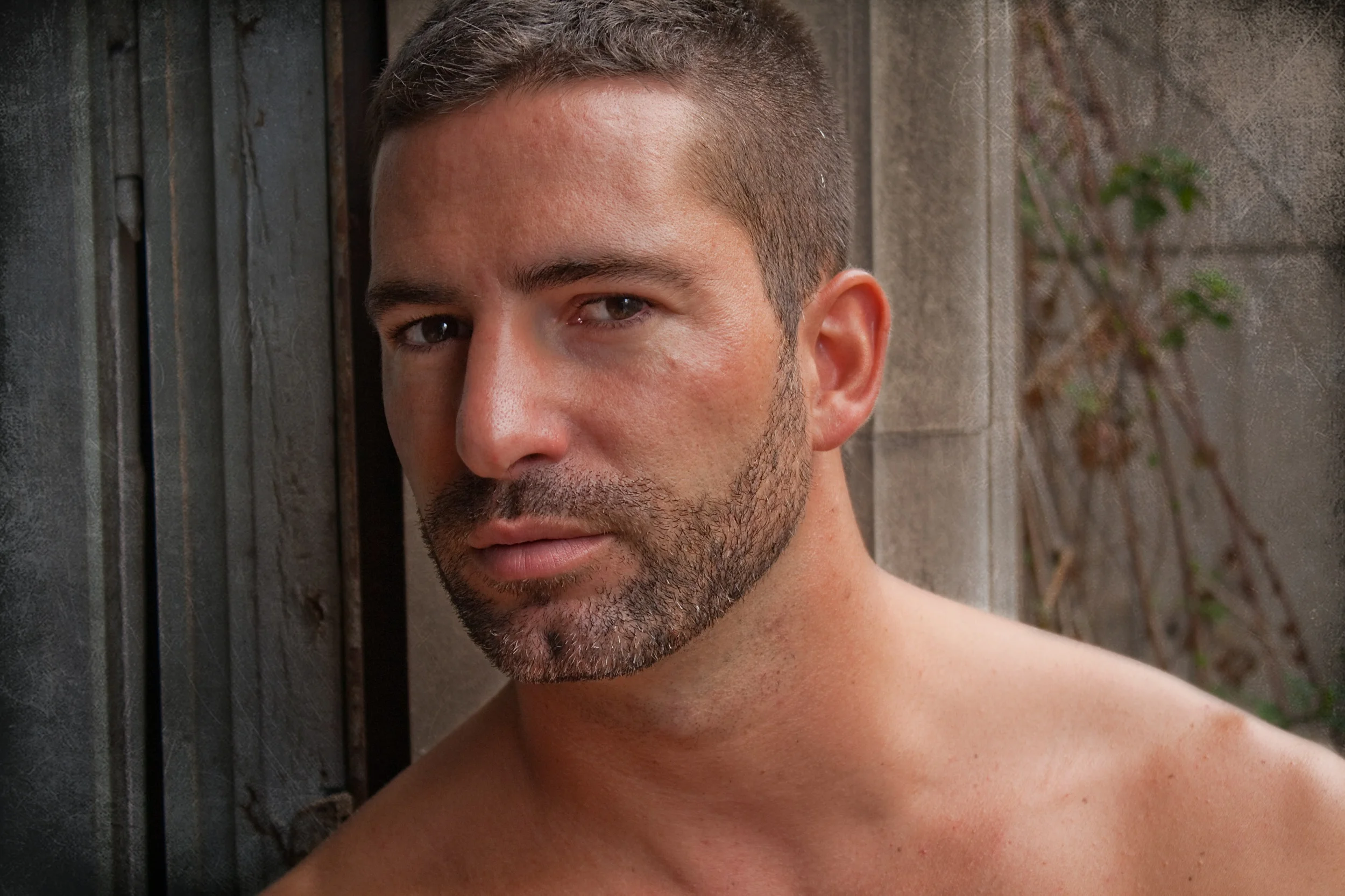 A shirtless man with short brown hair and a beard, leaning against a weathered wooden and concrete wall outdoors.