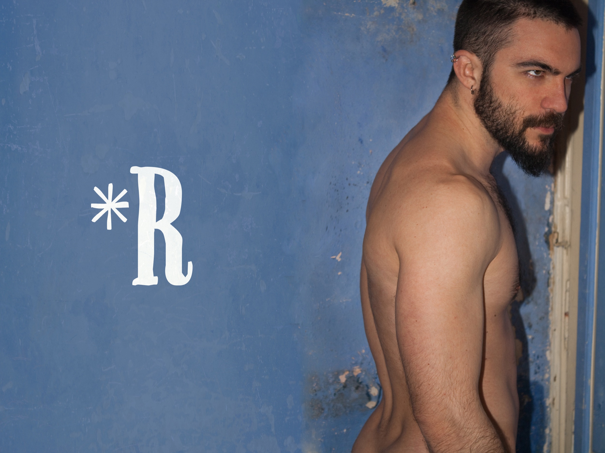 A shirtless man with dark hair and a beard, wearing an earring, standing against a distressed blue wall with the letter 'R' and an asterisk symbol on it.
