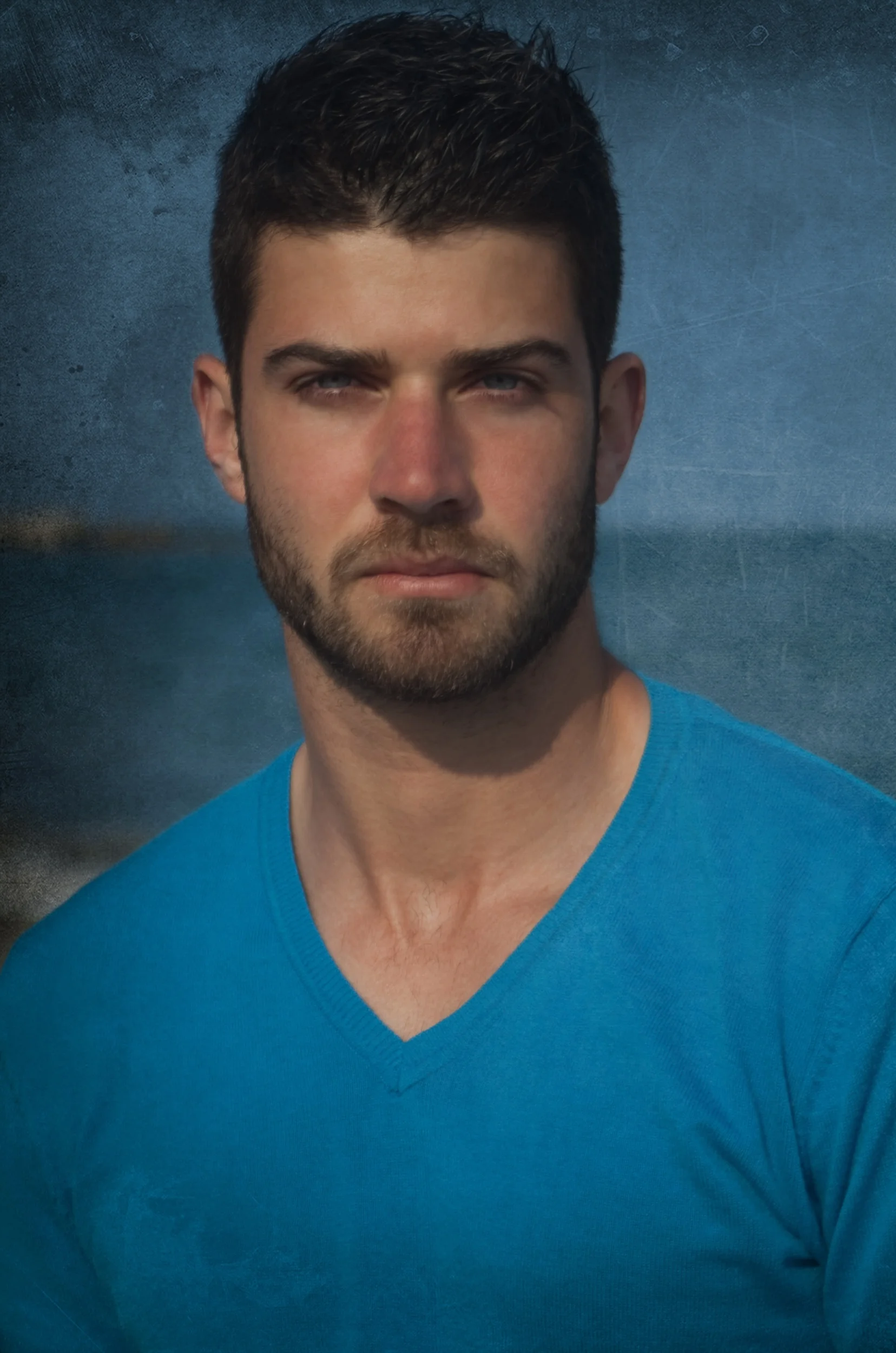 A young man with dark hair and a beard wearing a blue V-neck shirt, outdoors with a body of water and a cloudy sky in the background.