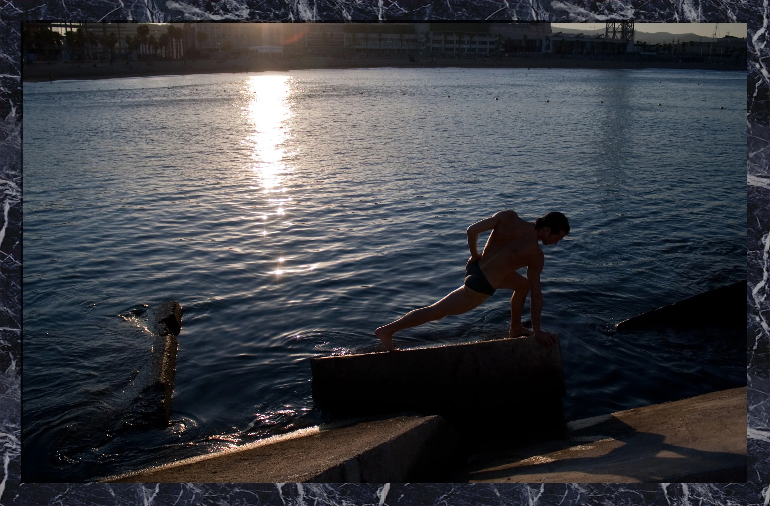 A shirtless man in swimming trunks is climbing onto a concrete ledge by the water at sunset, with the sun reflecting off the water's surface.