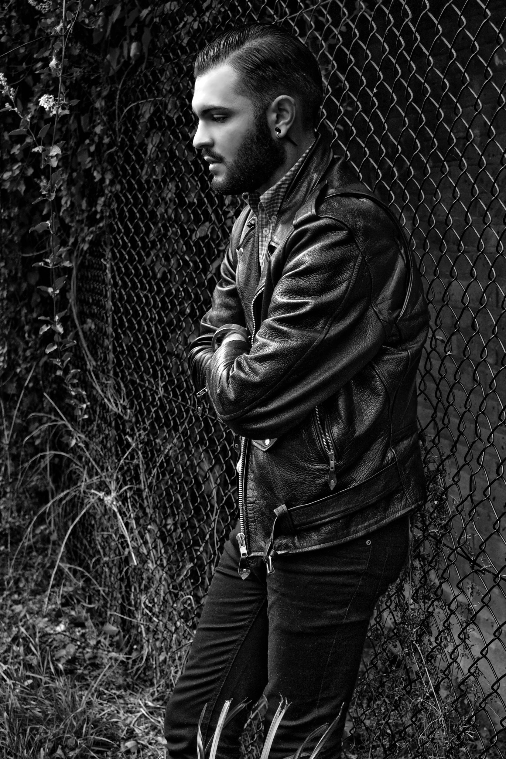 Black and white photo of a bearded man with slicked-back hair, wearing a leather jacket and a checkered shirt, leaning against a chain-link fence with plants growing around it.